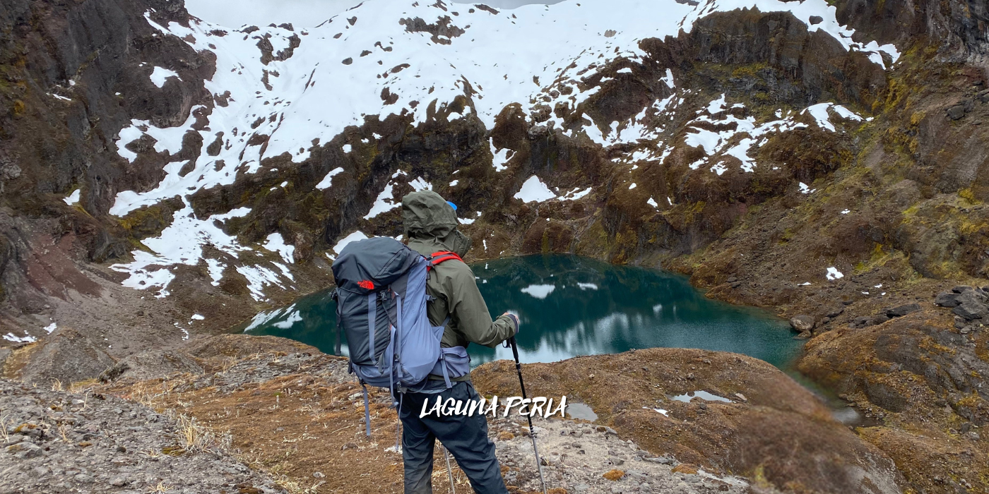 persona con equipo de senderismo frente a un lago rodeado de montañas con nieve