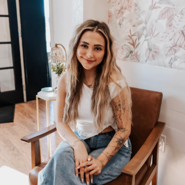 A young woman with long, curly brunette hair and a septum piercing, smiling at the camera, sitting on a wooden bench in front of a light patterned wall.