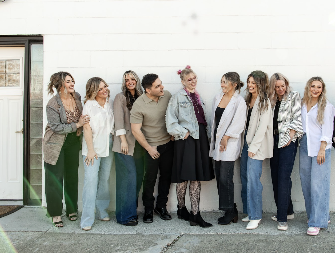 Group of nine women and one man standing against a white wall outdoors, smiling and laughing.