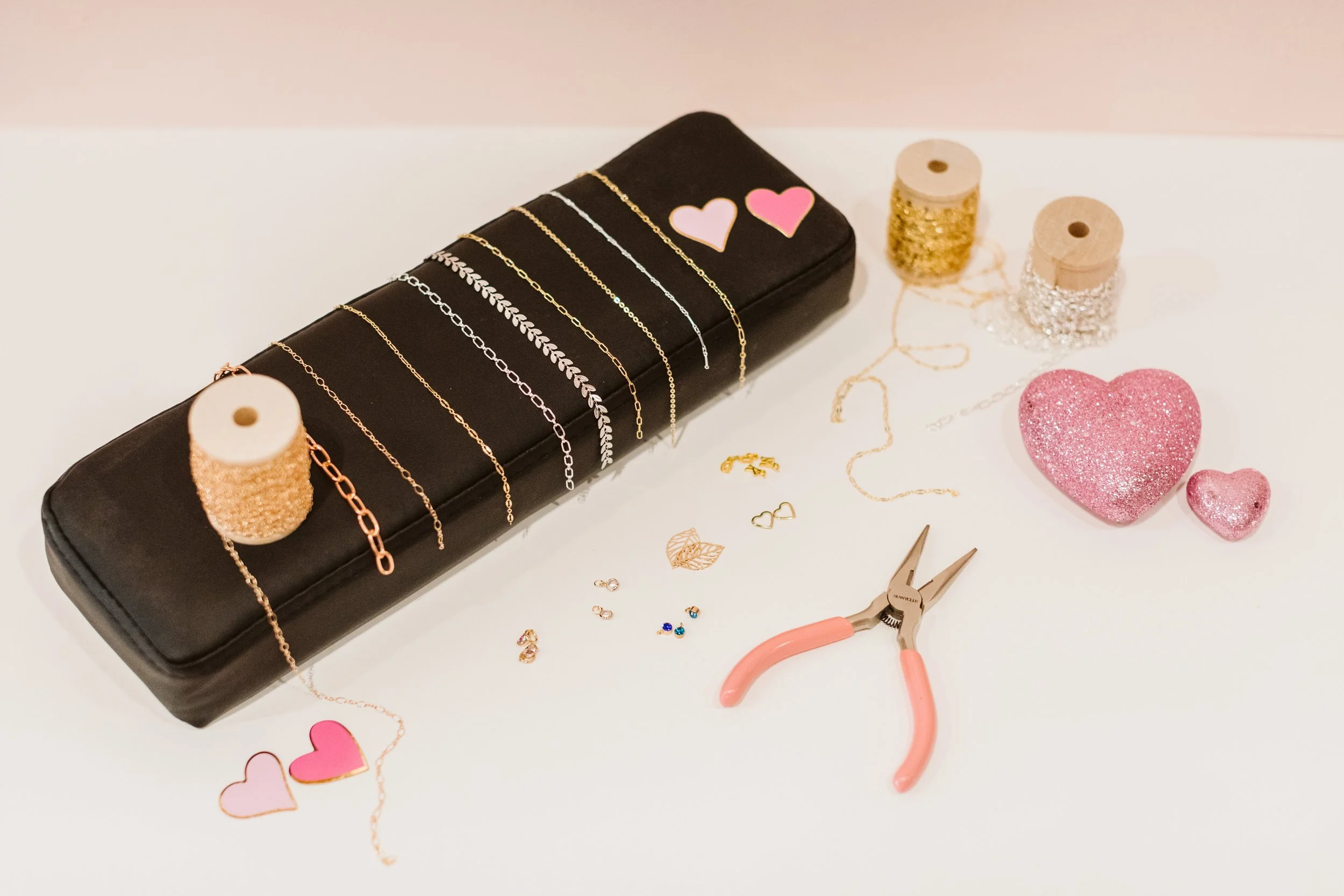 Jewelry-making workspace with a black case holding various necklaces, gold and silver thread spools, two pink glitter hearts, small jewelry charms, pliers, and paper heart cutouts.