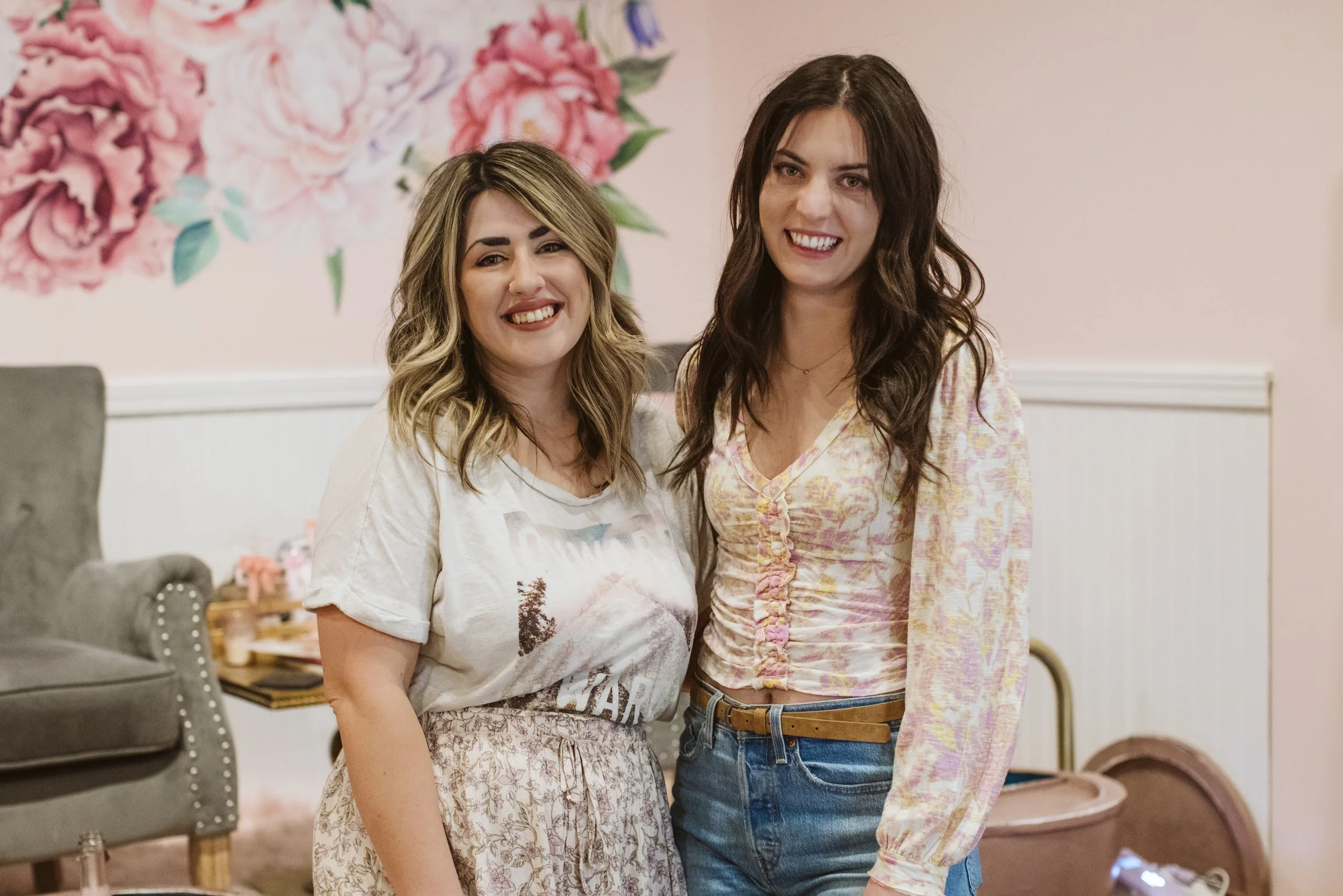 Two young women standing inside a room, smiling at the camera. One has blonde hair with darker roots and wears a graphic T-shirt and a patterned skirt. The other has dark brown hair and wears a colorful, patterned blouse with blue jeans. In the background, there is a pink wall with floral art, a grey chair, a small table with decoration, and some children's toys on the floor.