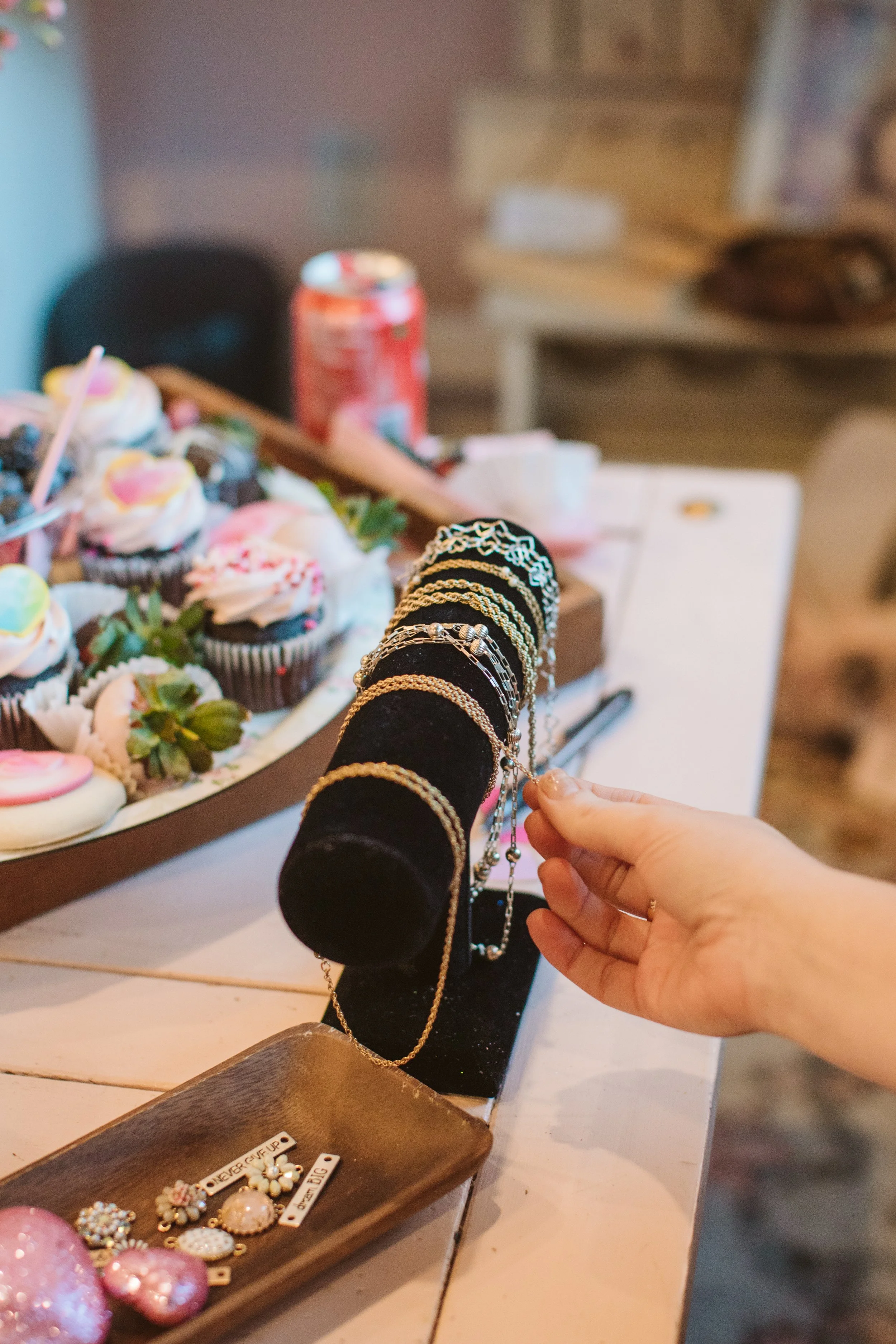A person's hand holding a necklace while browsing display jewelry on black velvet necklace holders, with a tray of earrings and nails painted pink in the foreground, and a plate of cupcakes and other treats in the background.