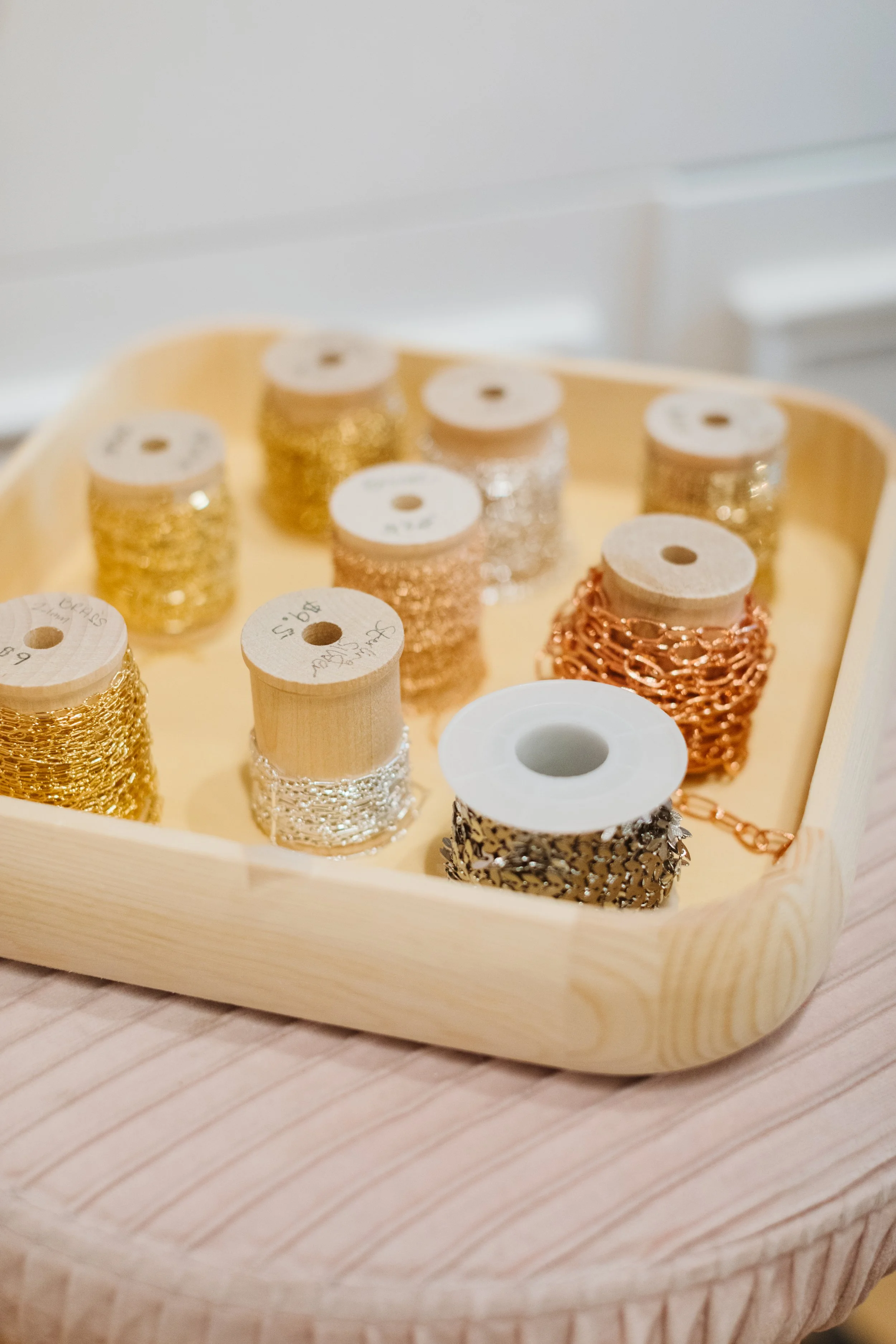 Wooden tray holding spools of decorative metallic chains in gold, silver, and rose gold colors.