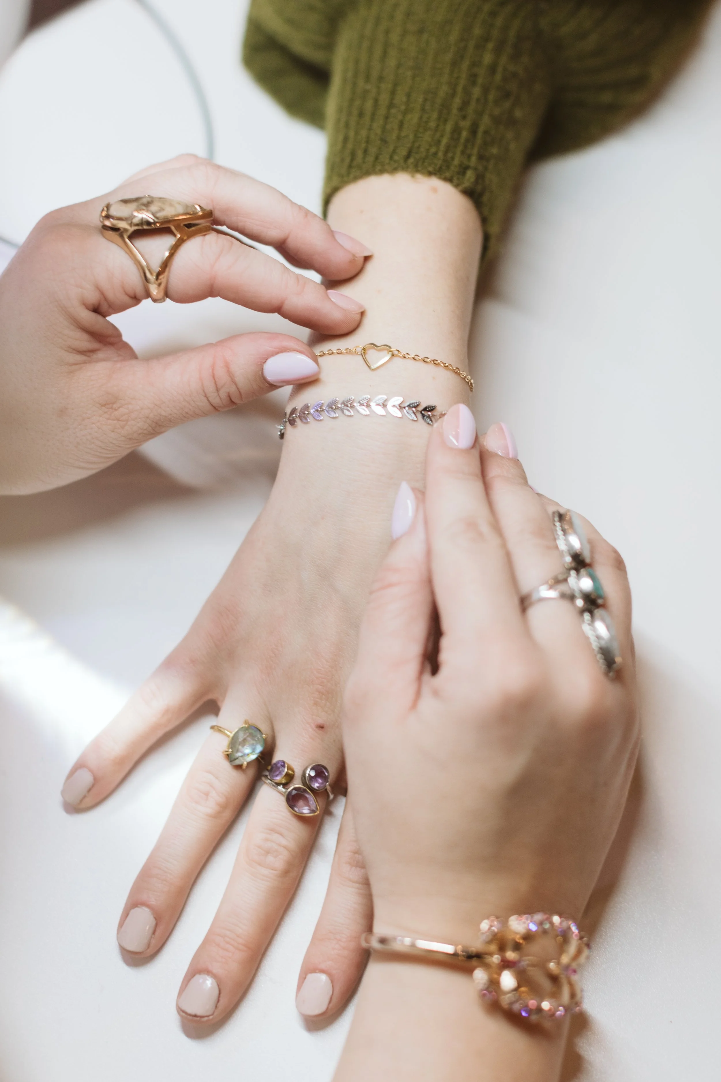 A person wearing multiple rings, bracelets, and a delicate chain with a heart pendant, while another person helps with jewelry on their wrist.