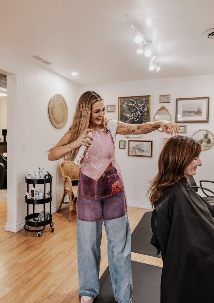 A hairstylist styling a woman's hair in a salon with artwork and a cart with hair products in the background.