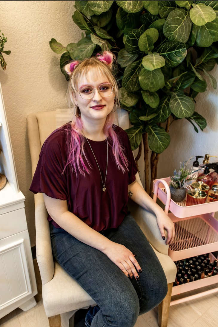 A young woman with pink hair styled in buns and long hair sitting on a beige chair near green plants. She is wearing glasses, a burgundy top, and dark jeans, with makeup and jewelry. There are beauty products on a pink trolley beside her.