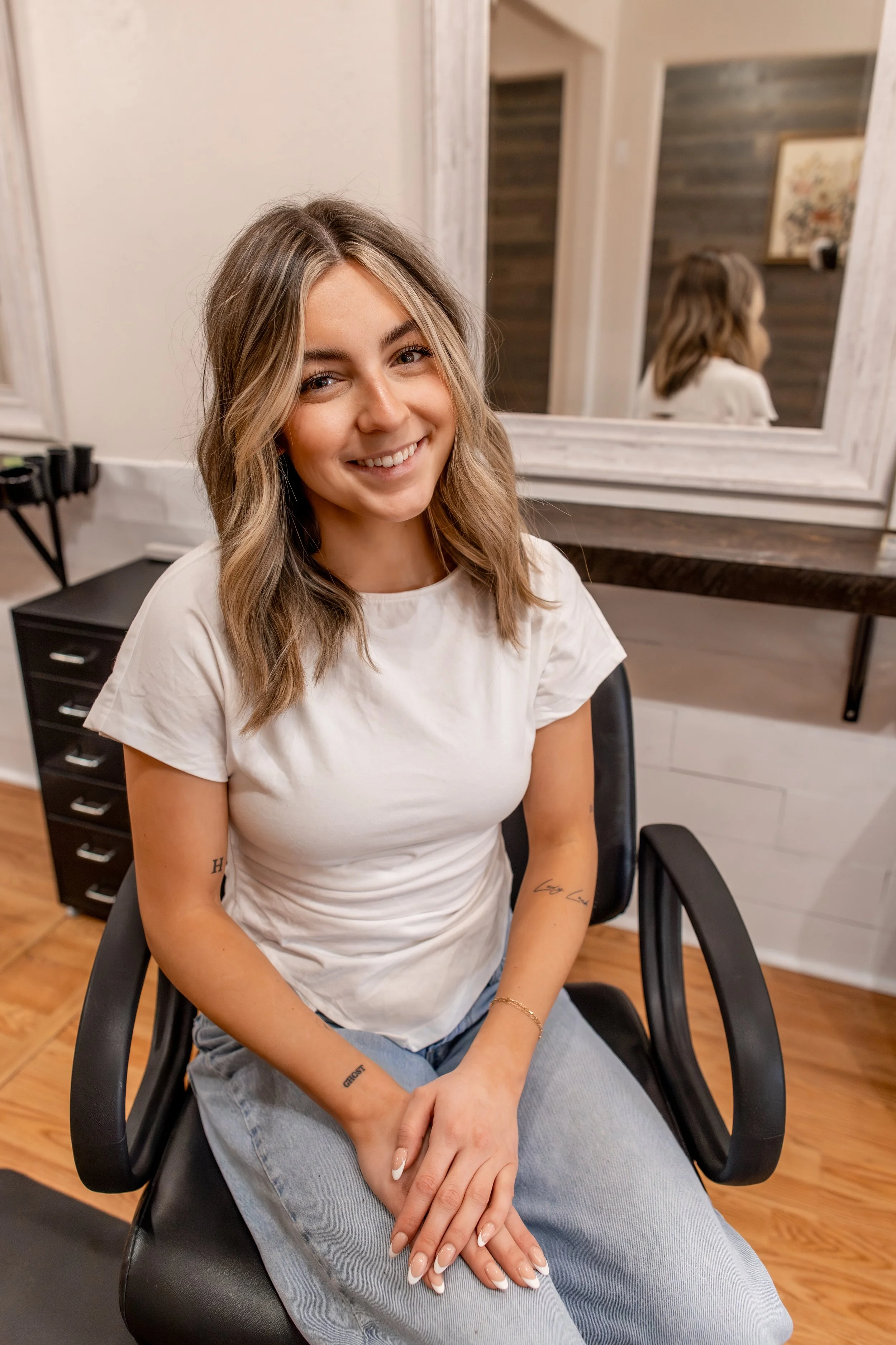 A young woman with long, wavy brown hair and light skin, smiling while sitting on a wooden chair with a rattan backrest in a bright, softly lit room.