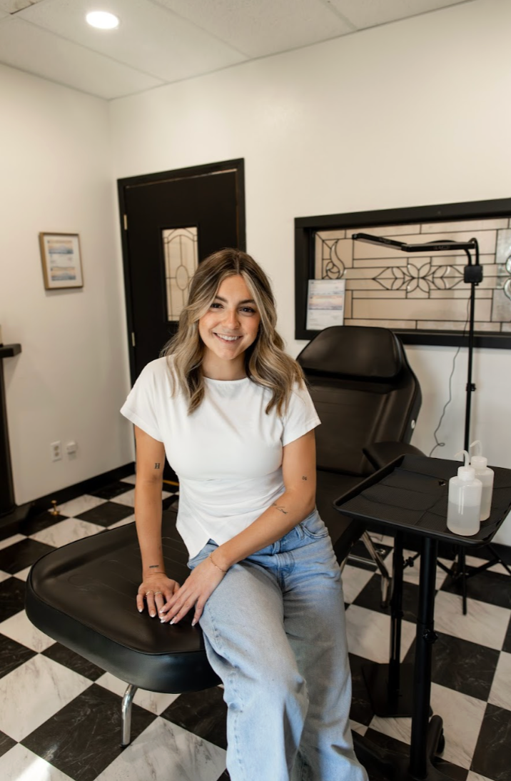 A young woman smiling and sitting on a black medical examination chair in a clinic or spa room with white walls, black and white checkered floor, and medical supplies on a small black table.