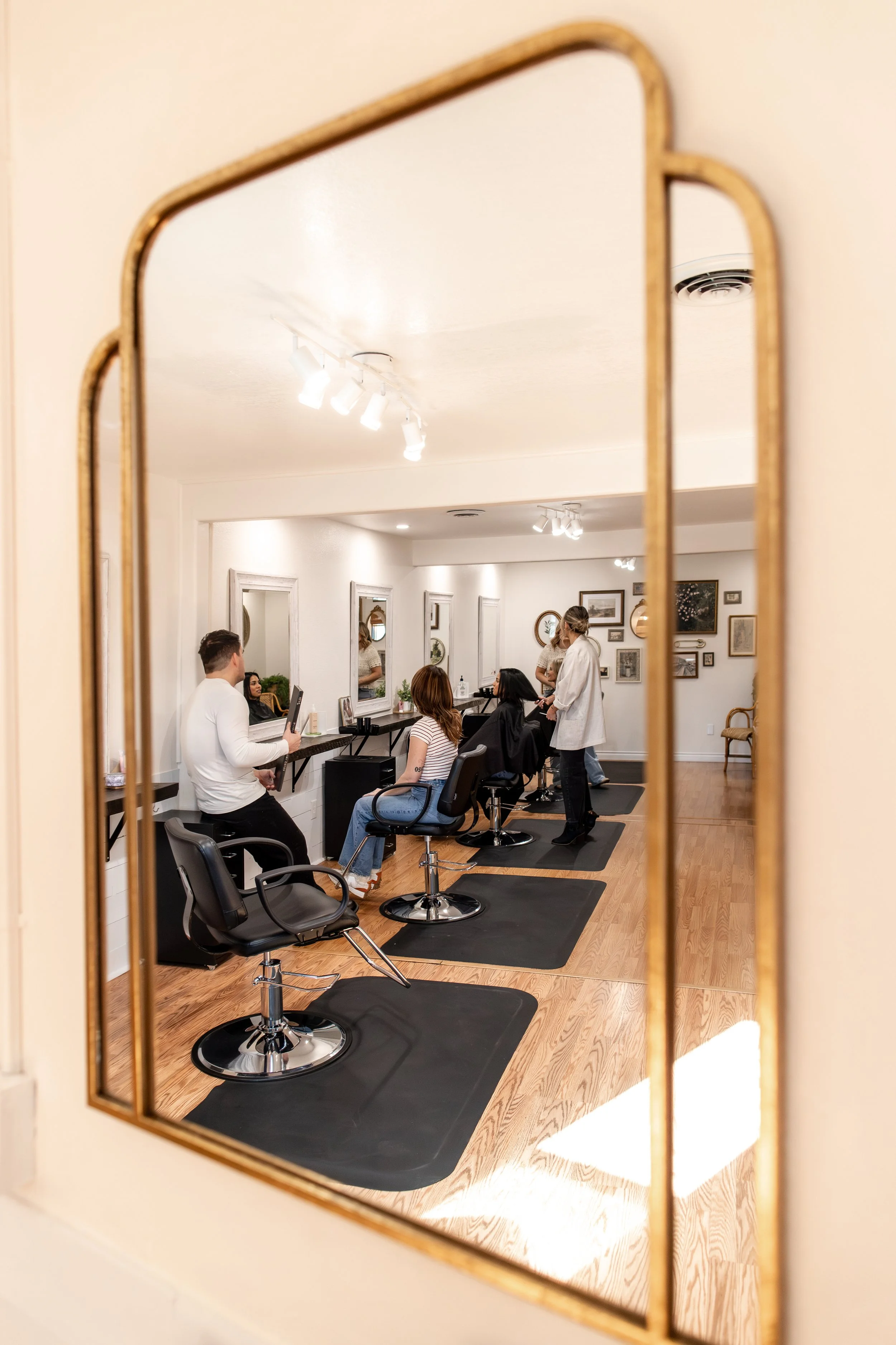 Beauty salon with hairstylist cutting hair of a client while another client sits waiting, reflection in a mirror, framed in a decorative mirror, with wall art and framed pictures in the background, wood flooring, ceiling lights.