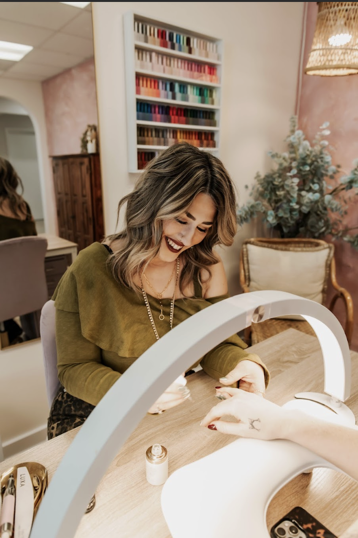 A woman with wavy blonde hair wearing a floral black top and pearl necklace smiling while getting a manicure.