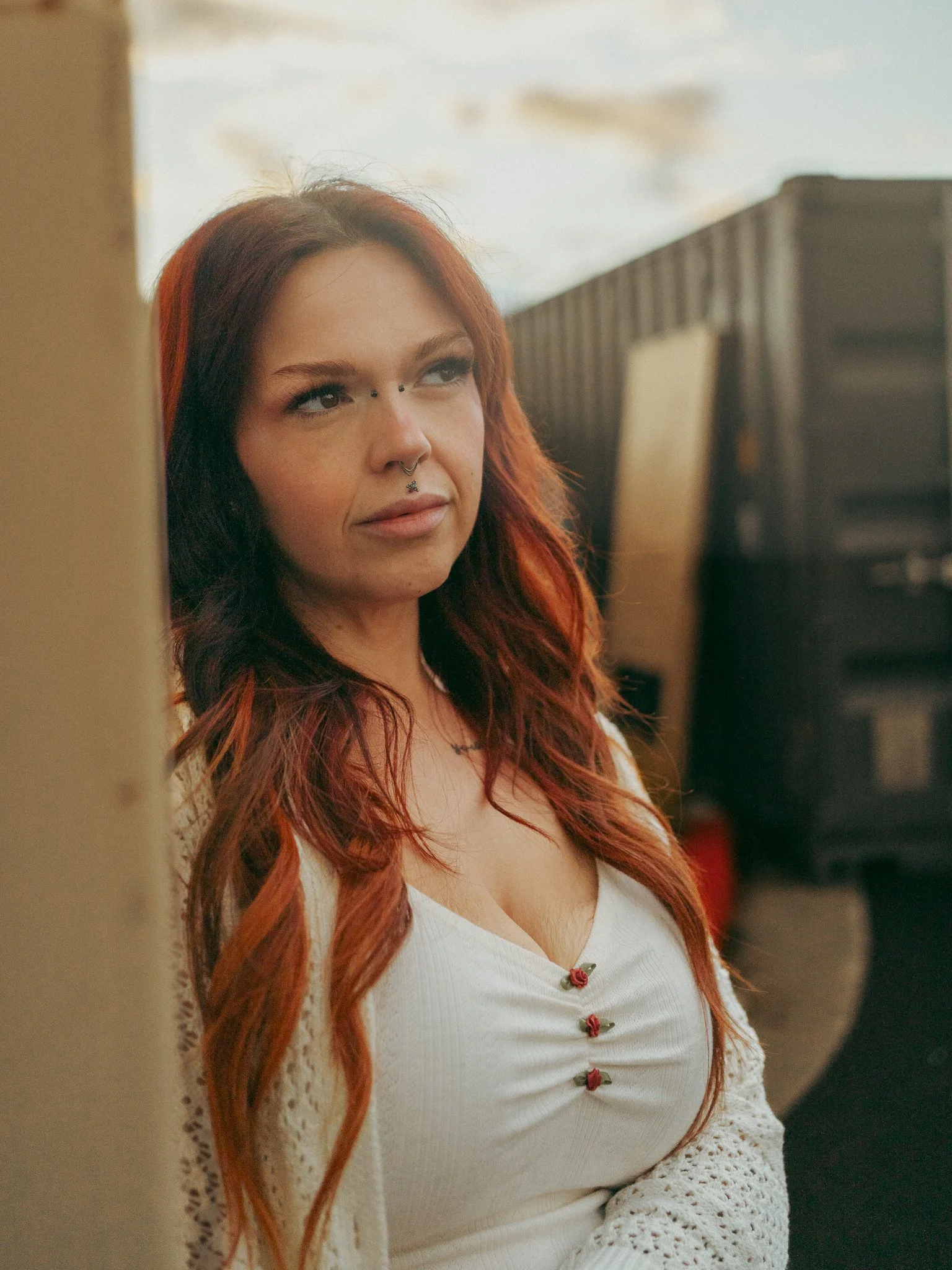 A woman with long red hair and multiple facial piercings looks to the side, standing outdoors near a beige wall with a trailer and sky in the background.