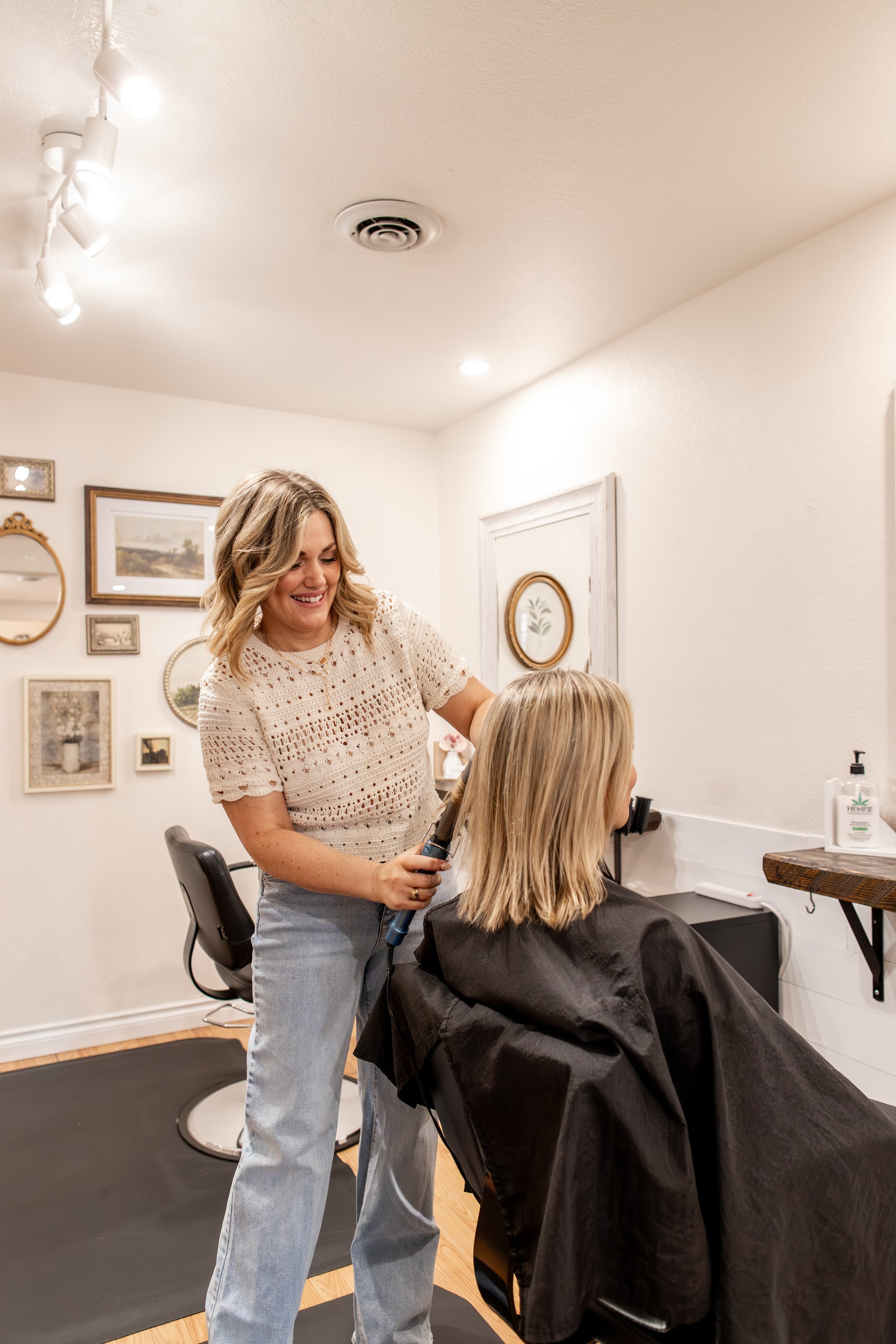 A hairstylist with shoulder-length blonde hair and a rust-colored top is cutting and styling a client's long, blonde, wavy hair in a salon with white decorative folding screens and a black shelf with a pink flower, hair products, and a mirror on the wall.