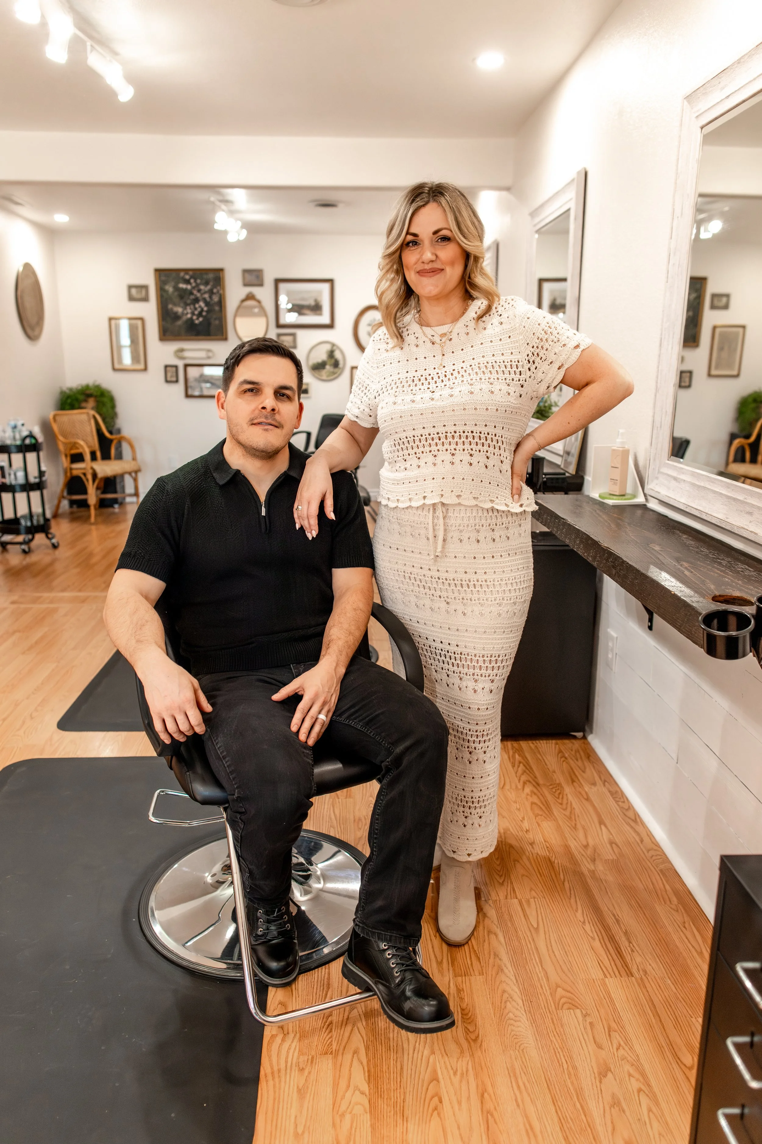A woman standing with her hand on her hip next to a man seated in a barber chair inside a salon.