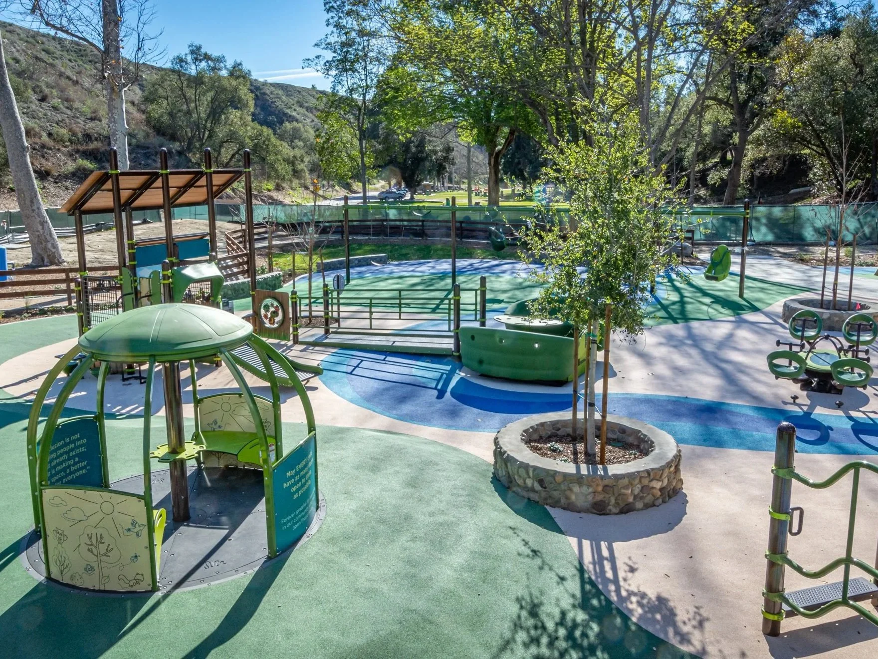 Ventura, California, children's playground with swings, a small round house, trees, and a pathway, surrounded by a green fence and trees with hills and parked cars in the background.