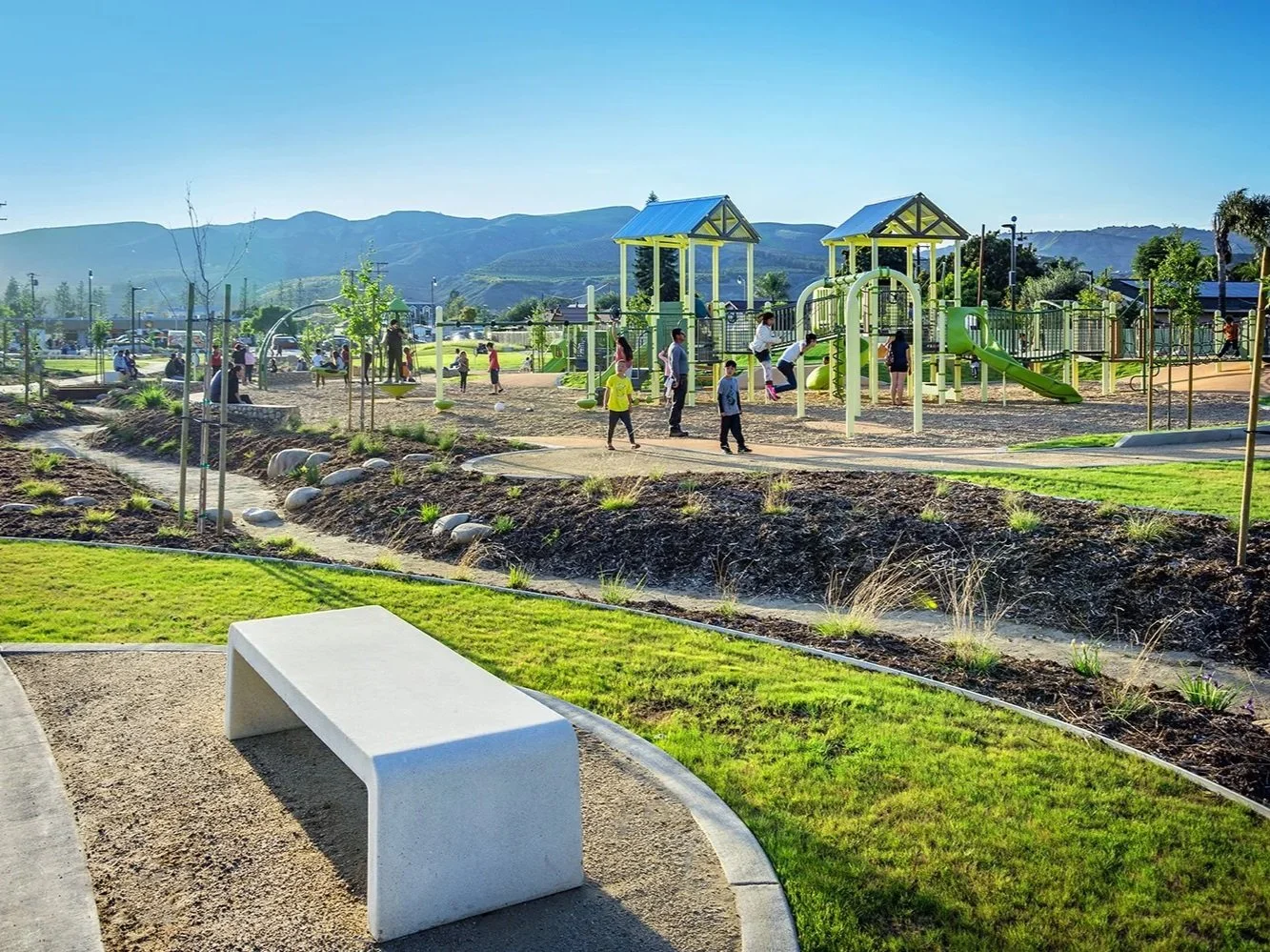 Ventura park with playground equipment including slides and climbing structures, children and adults enjoying the area with mountains in the background, green grass, trees, and a white bench in the foreground.