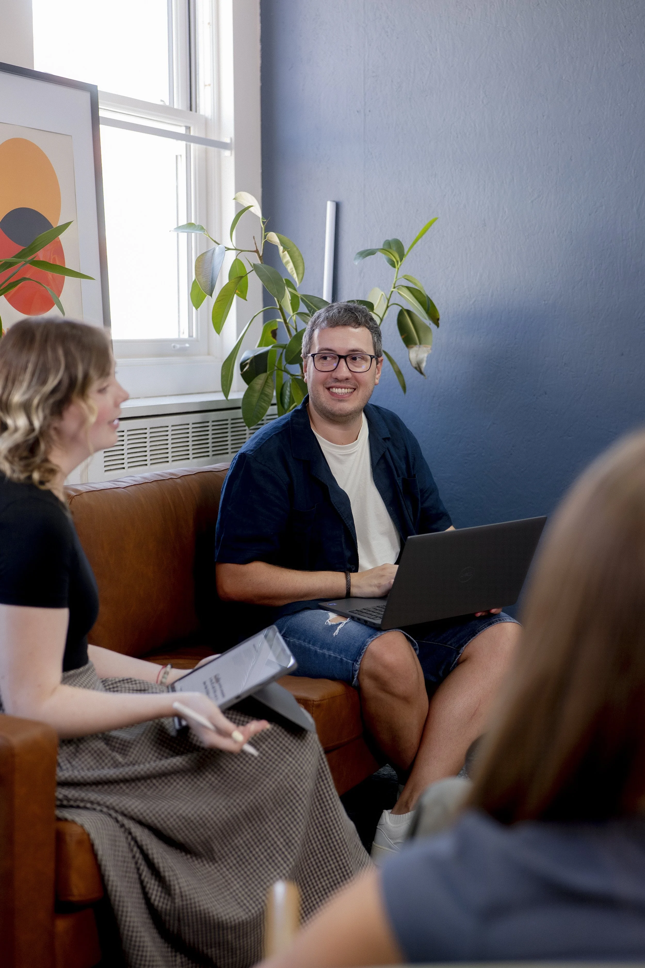 man smiling at coworkers wearing a white t-shirt and blue over shirt, with laptop on his lap.