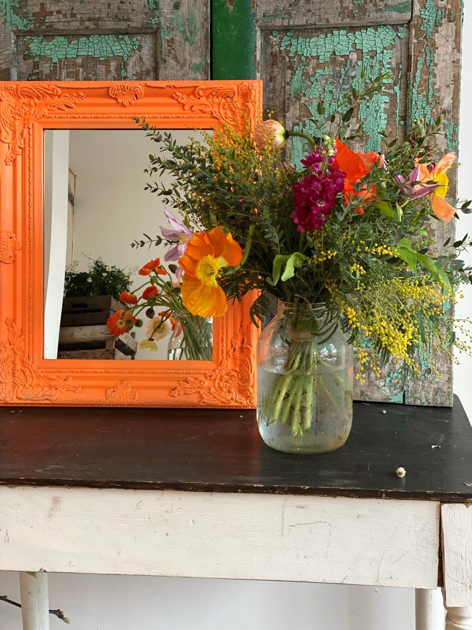 A brightly coloured vase arranged by a floristry basics student,  placed on a wooden table with a fluro orange mirror and old bottled green shutters