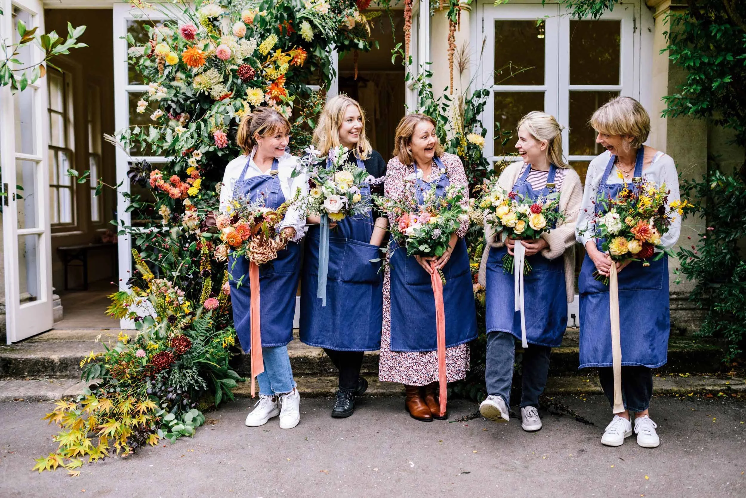 Five women standing outdoors in front of a floral backdrop, holding bouquets and wearing blue aprons. They are smiling and engaging with each other, surrounded by greenery and flowers, with a house and open door behind them.