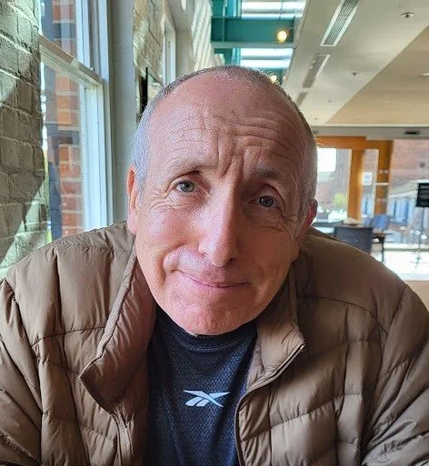 Close-up of a middle-aged man with a slight smile, wearing a beige puffer jacket and black shirt, sitting indoors with large windows and a modern interior in the background.