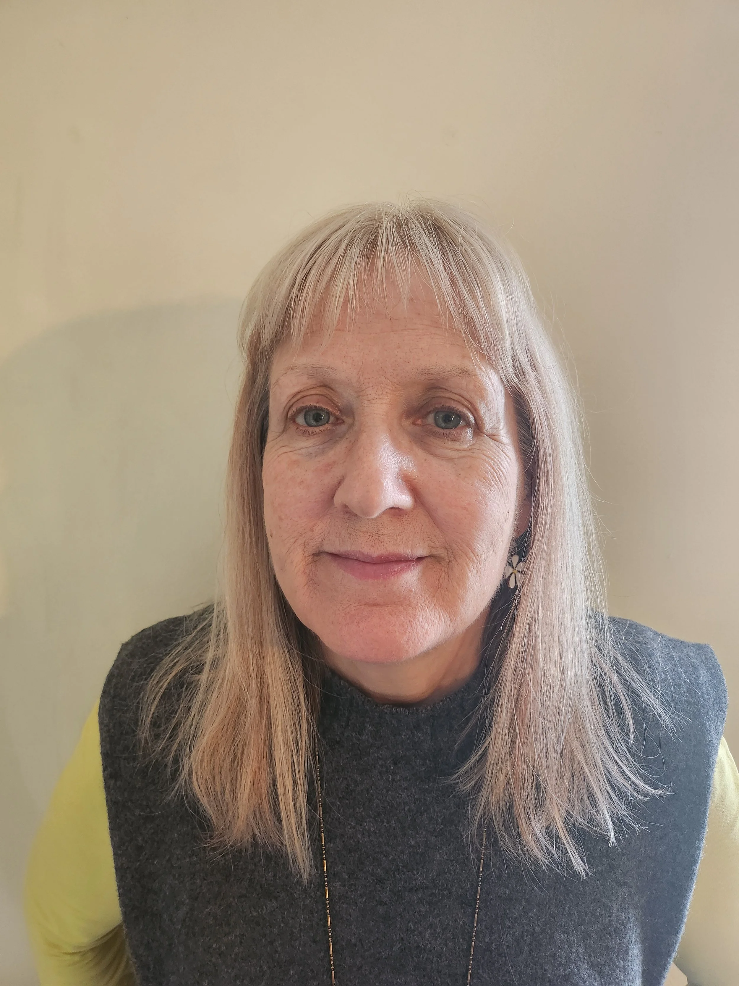 A middle-aged woman with shoulder-length blonde hair, blue eyes, and a neutral expression, wearing a gray sleeveless top over a yellow long-sleeve shirt and a subtle necklace, standing against a plain light-colored wall.