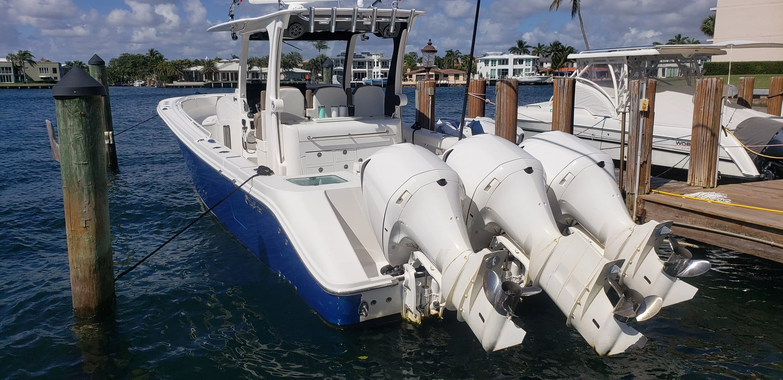 White fishing boat with three outboard motors docked at a marina, with houses and palm trees in the background.