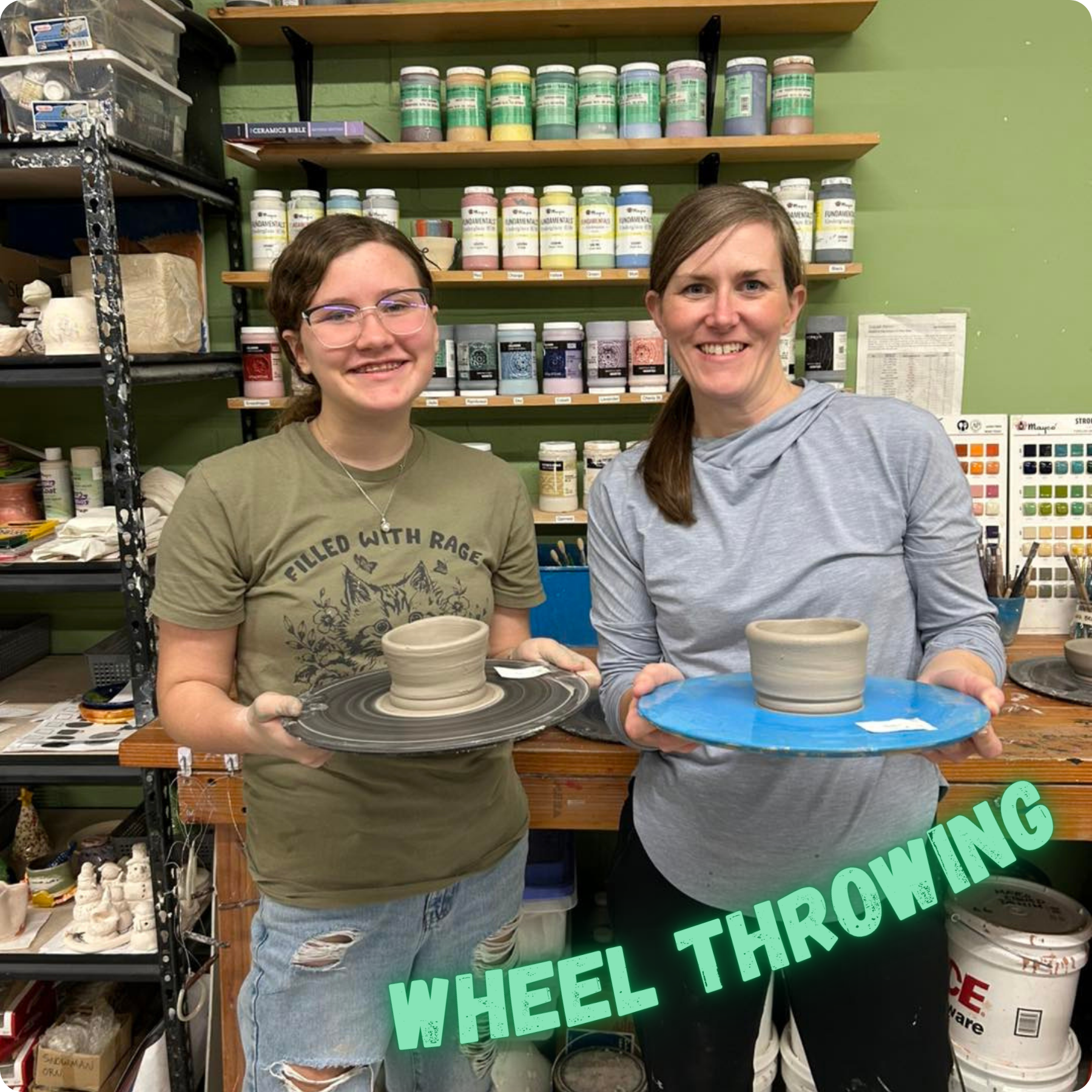 Two women smiling while holding pottery wheels and ceramic pieces in a ceramic studio, with shelves of paints and supplies in the background.