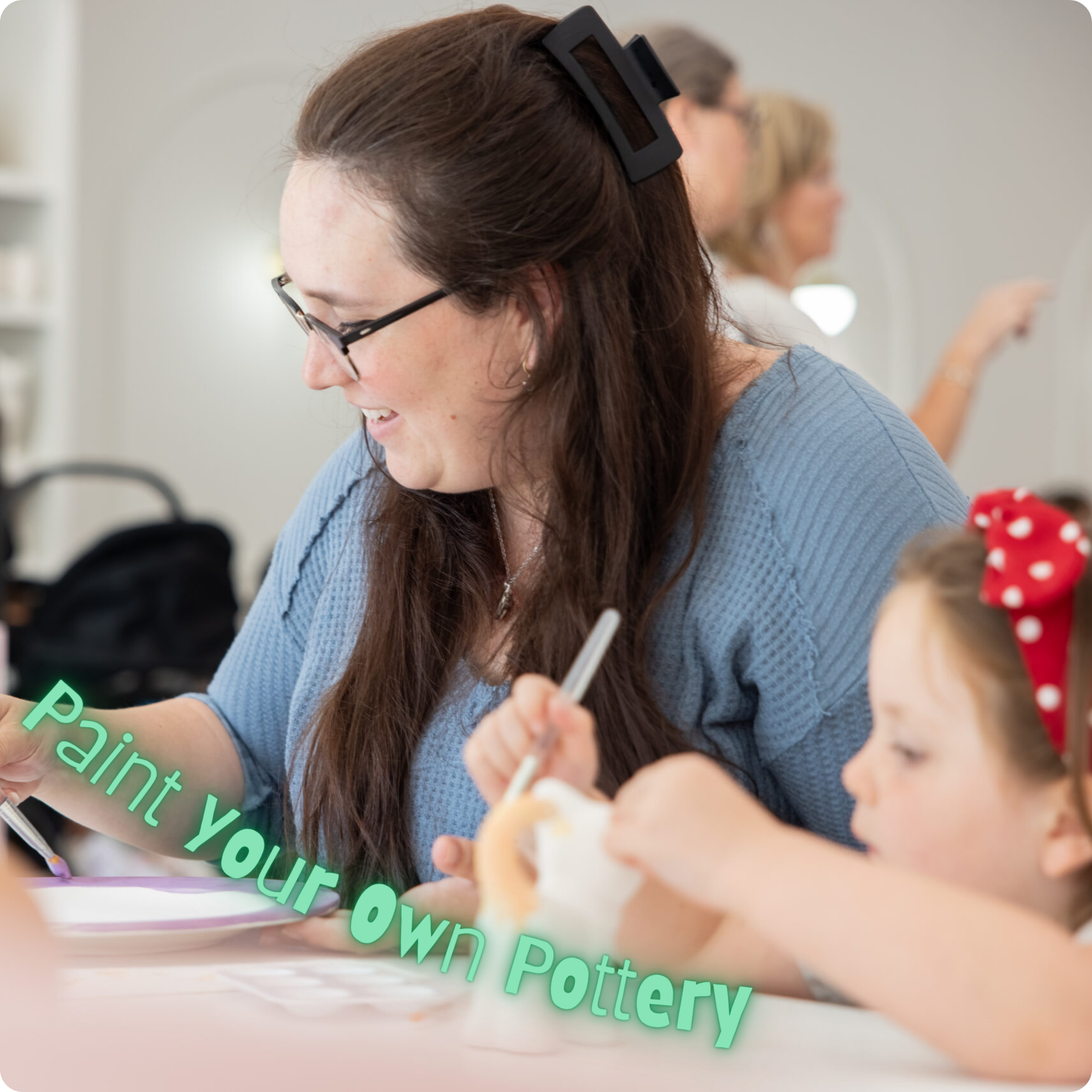 A woman with long brown hair and glasses, smiling and writing at a table, with children and other adults in the background, participating in a pottery activity.