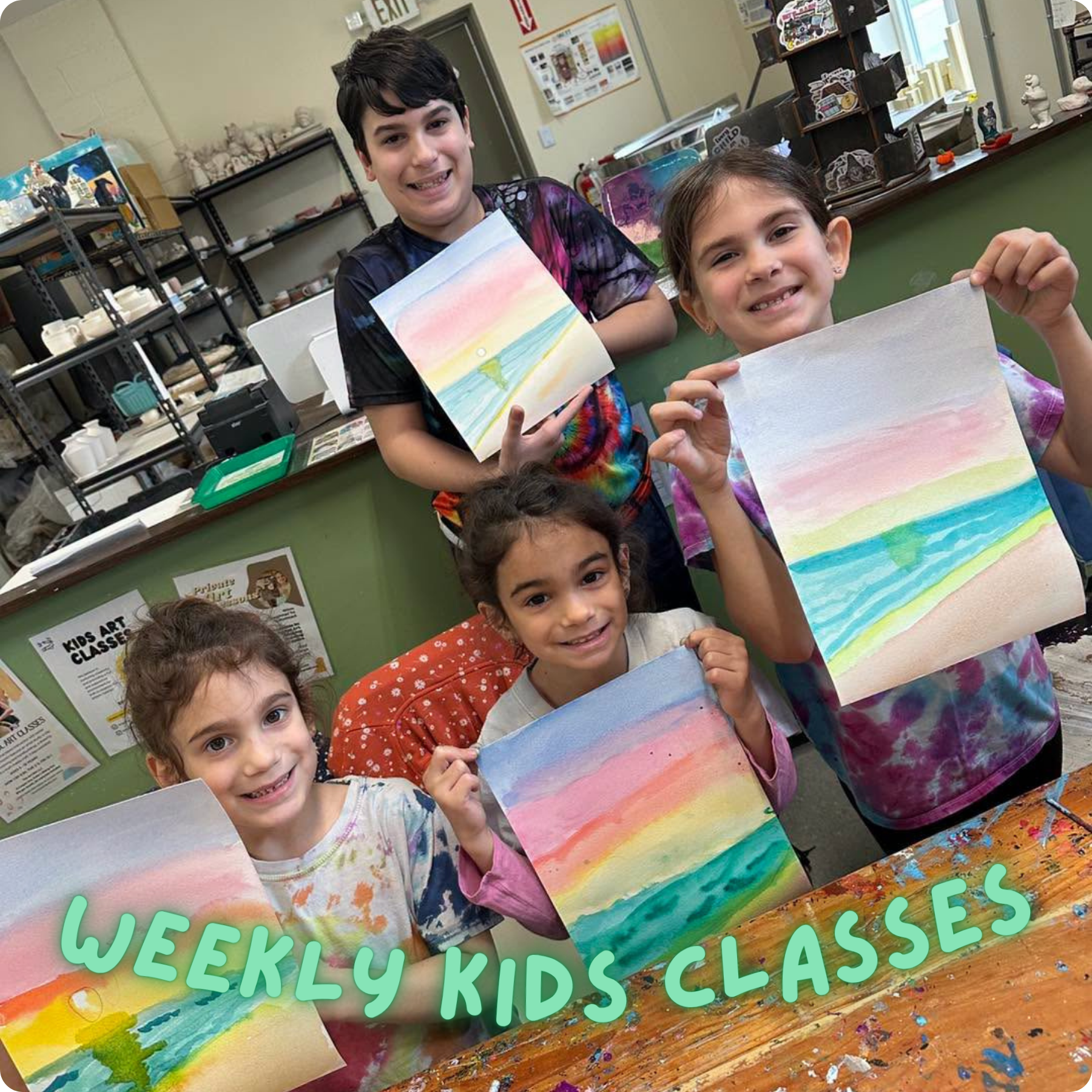 Children in an art classroom holding colorful watercolor paintings of a sunset over the ocean, with the message 'Weekly Kids Classes' written across the photo.