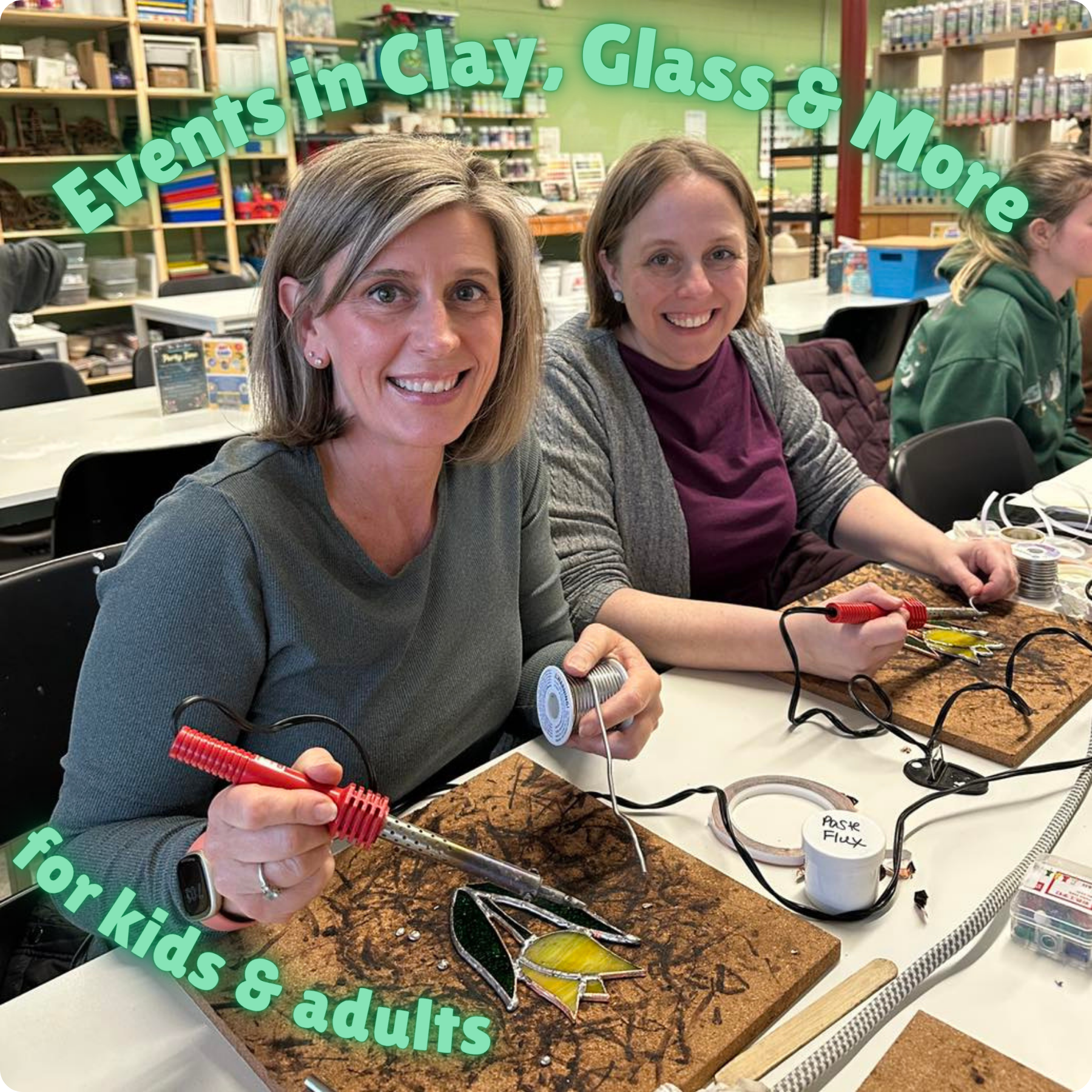 Two women are sitting at a table working on stained glass projects with tools and colorful glass pieces, in a craft room with shelves of art supplies in the background.