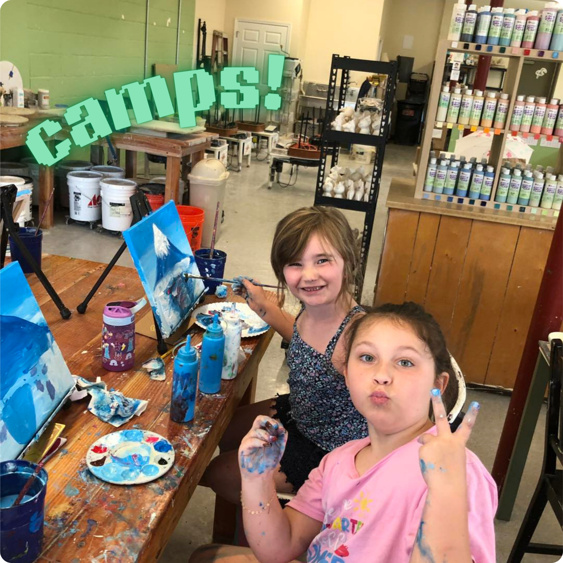 Children painting and smiling at a crafting table covered in paint supplies in an art studio.