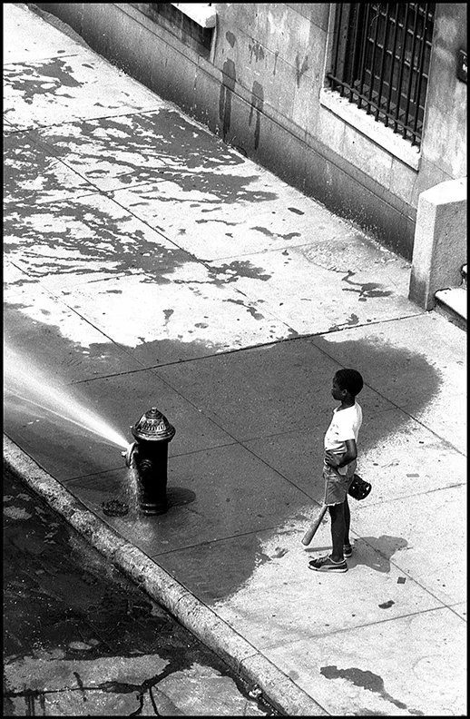 a young man, hot from a baseball game in Riverside Park, stopping to enjoy the cool water of an open hydrant. Drew Carolan, UWS, Manhattan [W. 88th St. on the UWS], July 1975.