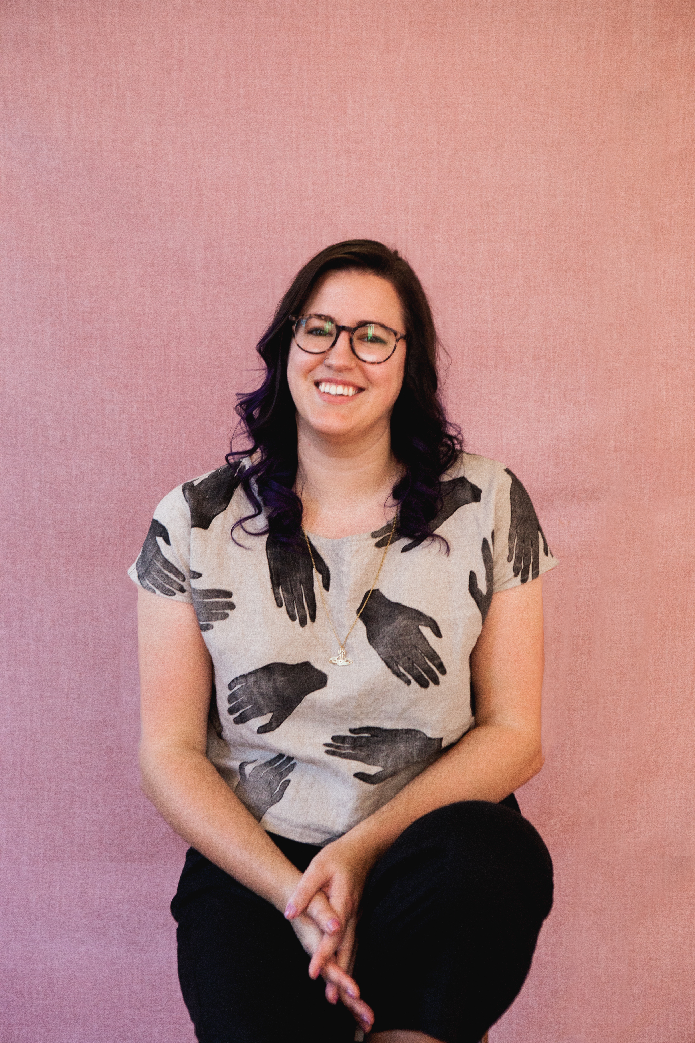 Professional headshot of NZ artist Nicole Keane sitting in front of a pink linen backdrop, wearing round black glasses and a tan linen shirt with black hand prints, smiling at the camera