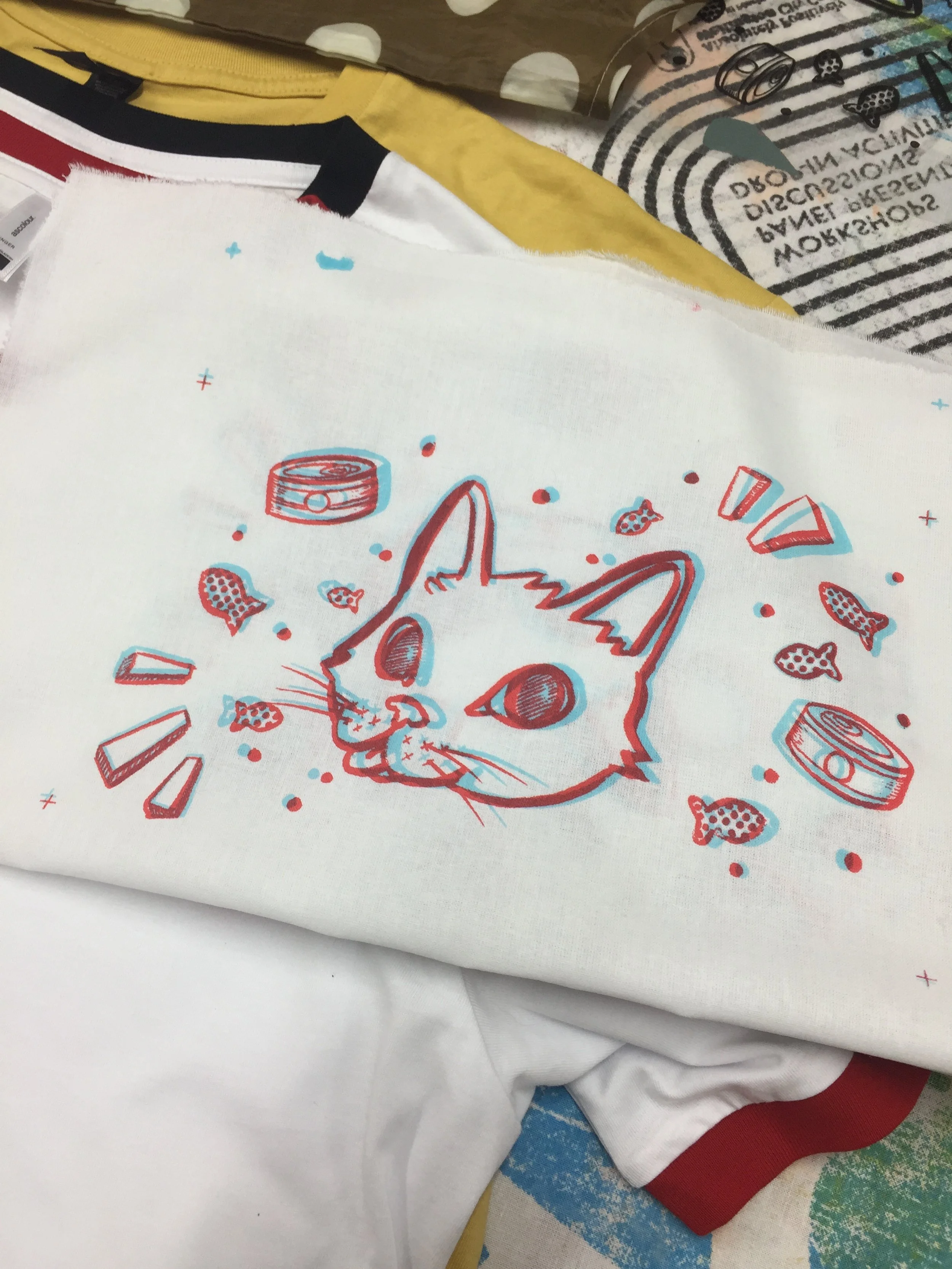 White t-shirt with retro 3D glasses at the collar, screen printed with a hand drawn cartoon cat head surrounded by fish and cat food cans in blue and red anaglyph style, by NZ artist Nicole Keane