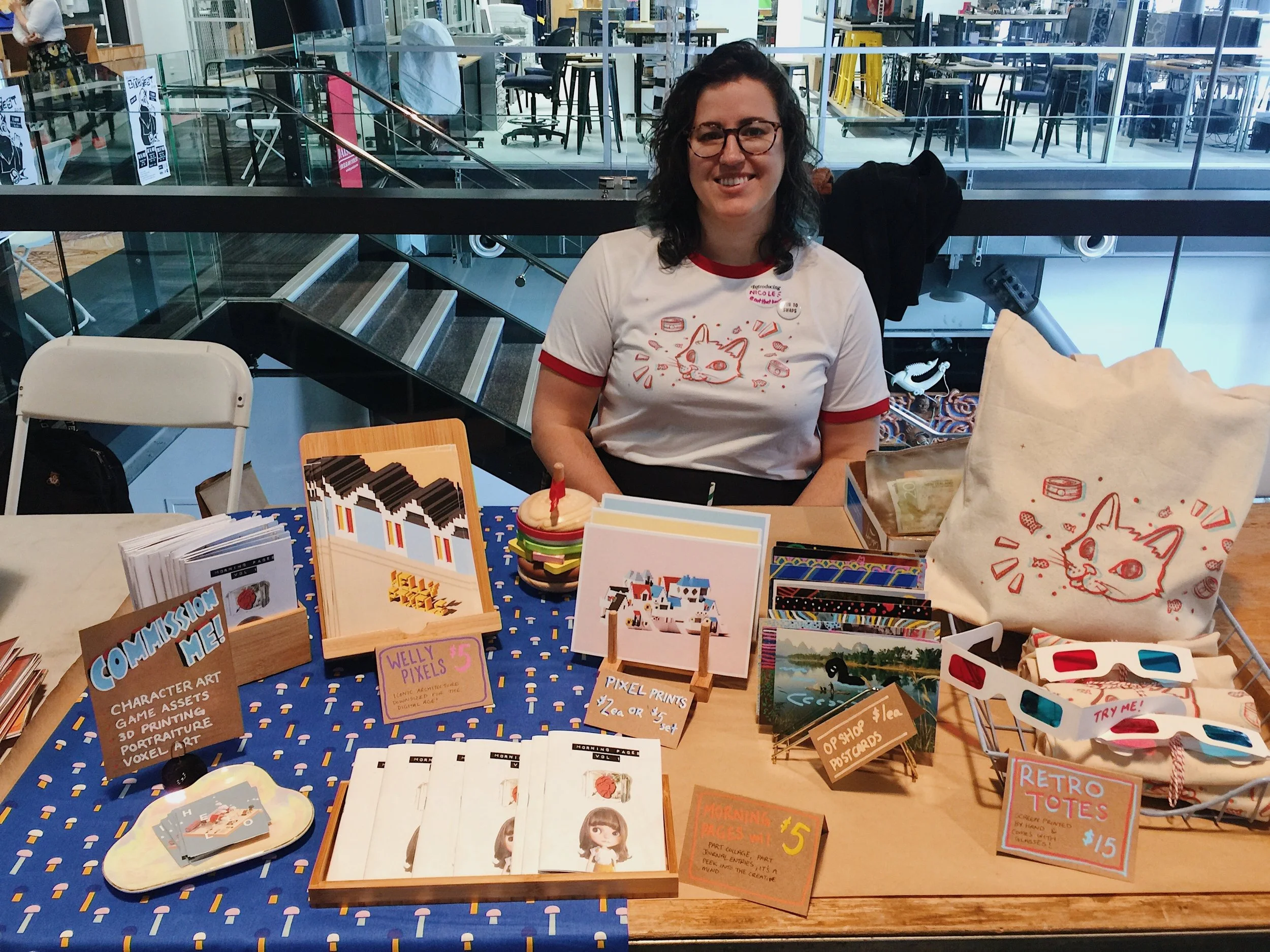 Nicole Keane smiling behind her stall at Wellington Zinefest 2019, displaying Morning Pages zine, Welly Pixels zine, pixel prints, anaglyph cat tote bags, a commission me sign, and Sleepy City business cards, wearing the anaglyph cat t-shirt