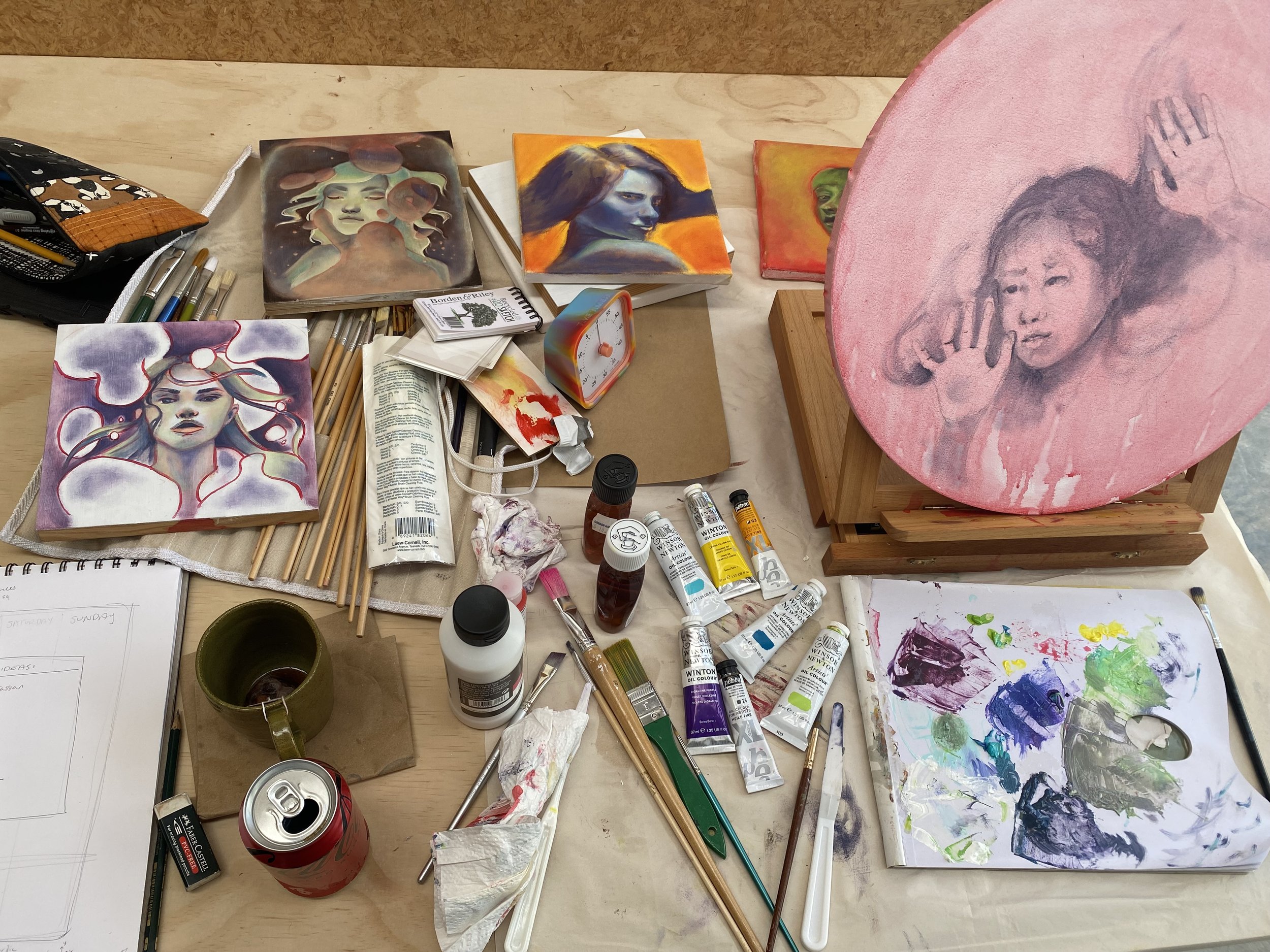 Nicole Keane's oil painting studio desk showing a circle canvas in progress on an easel, four paintings in various stages, brushes, oil paints, palette, sketchbook and half finished drinks