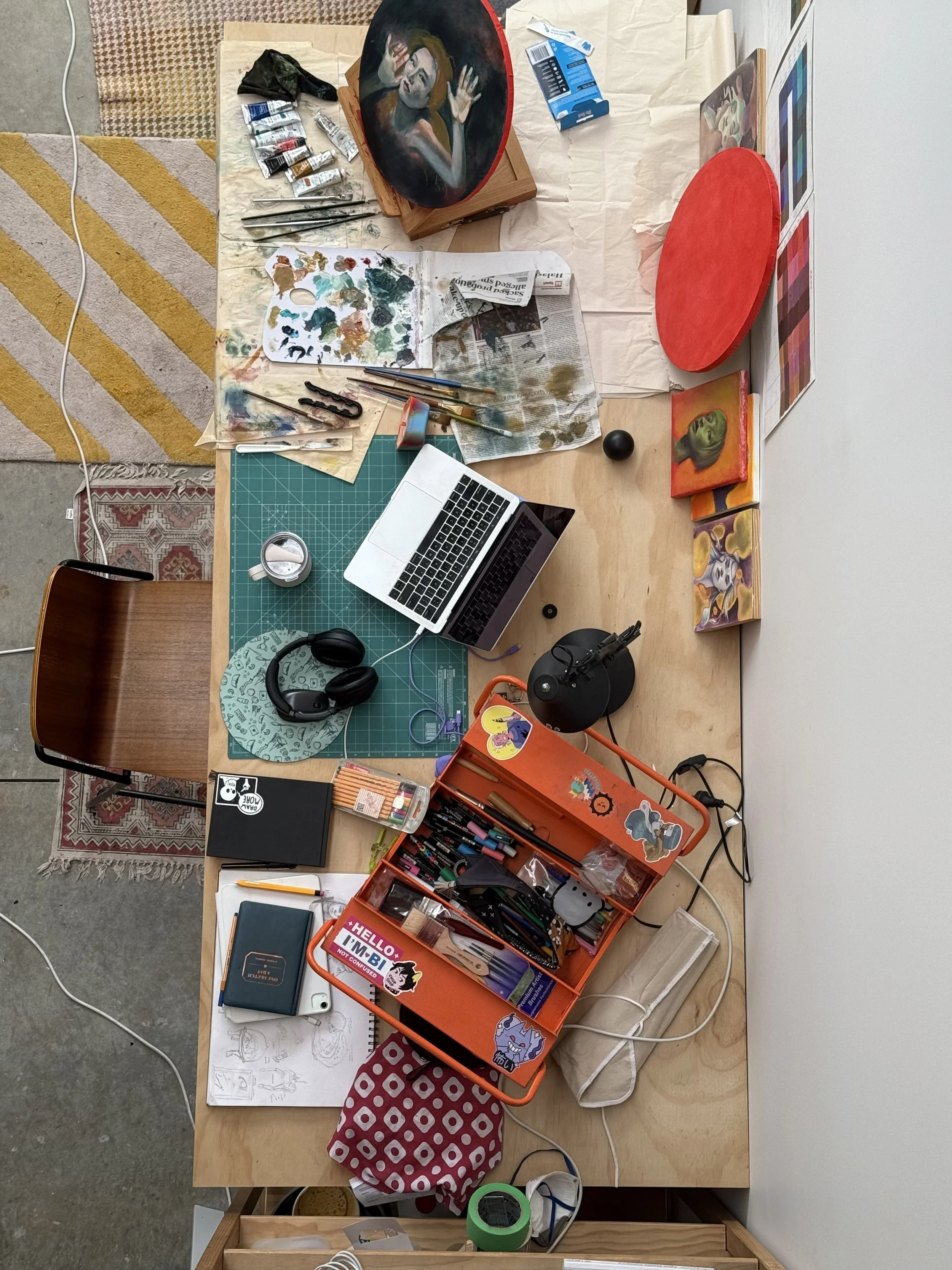 Birds eye view of NZ artist Nicole Keane's studio work table showing paintbrushes on newspaper, a green cutting mat, MacBook, sketchbook, chunky headphones, sticker covered toolbox with art supplies, and three paintings leaning against the wall