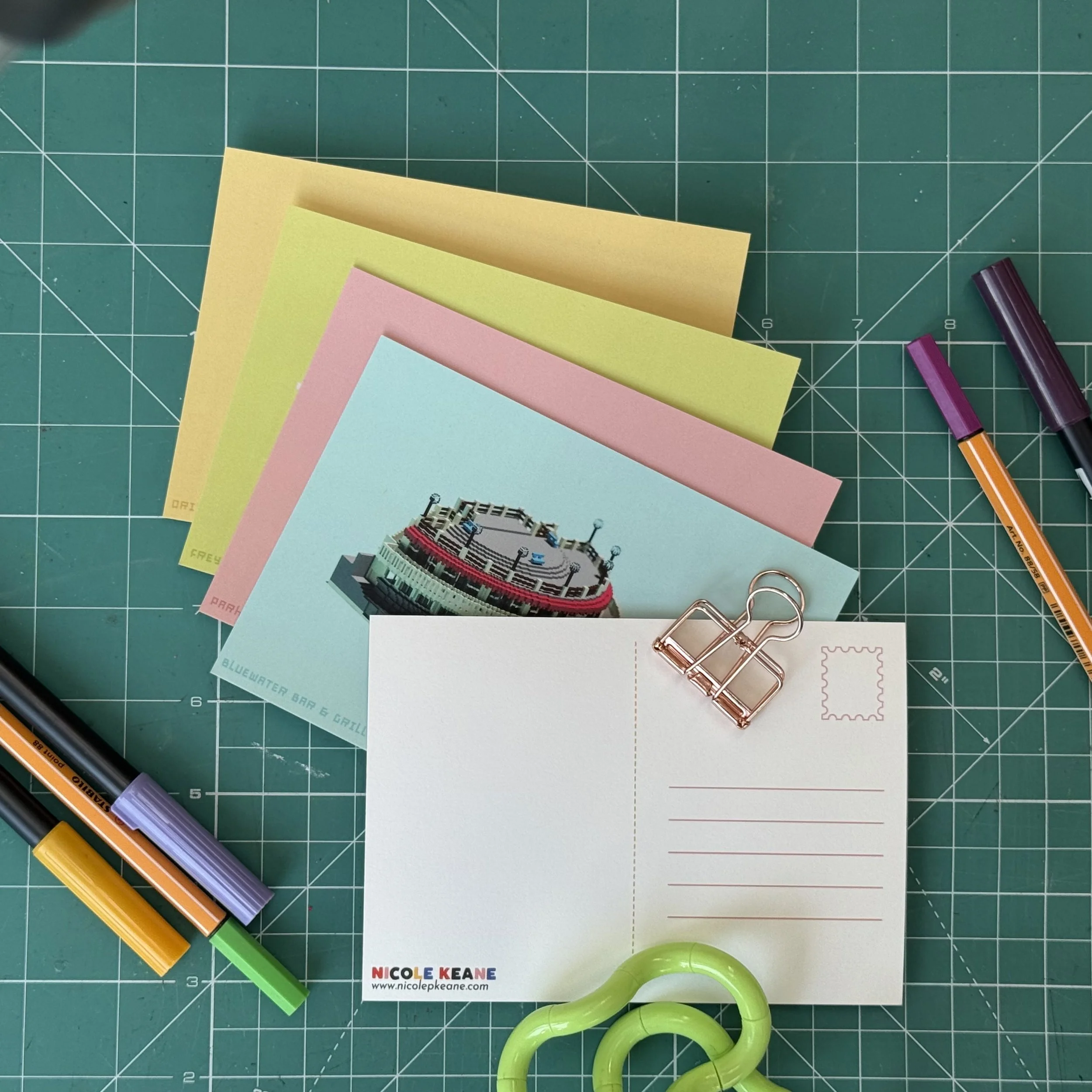 Flat lay of the Welly Pixels postcard set fanned out on a green cutting mat, showing pastel colours and the Rotunda design alongside the custom-designed back with address lines and stamp box