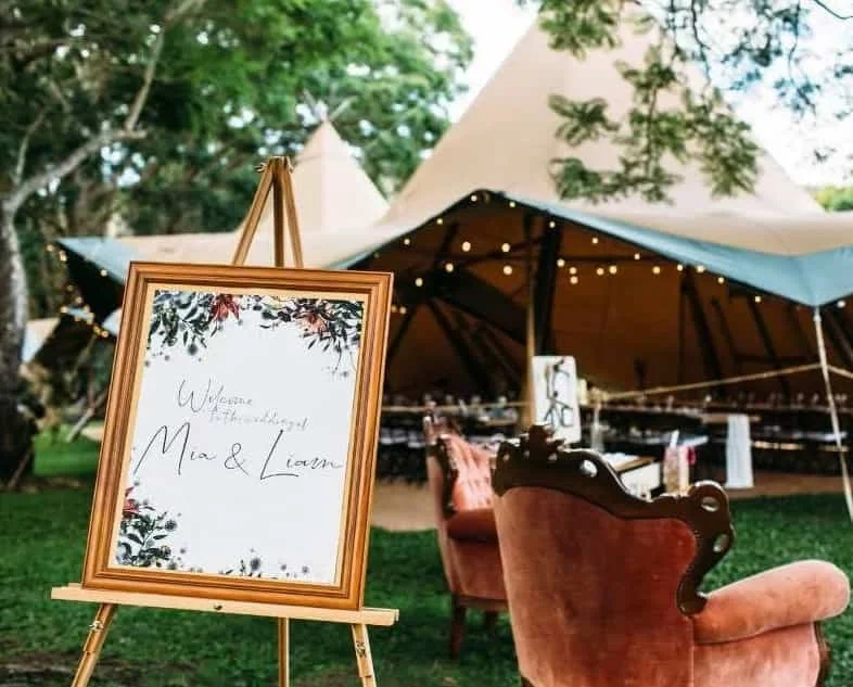 Wedding reception setup with a sign welcoming guests to Mad & Lauren, outdoor tent with string lights, chairs, and tables.