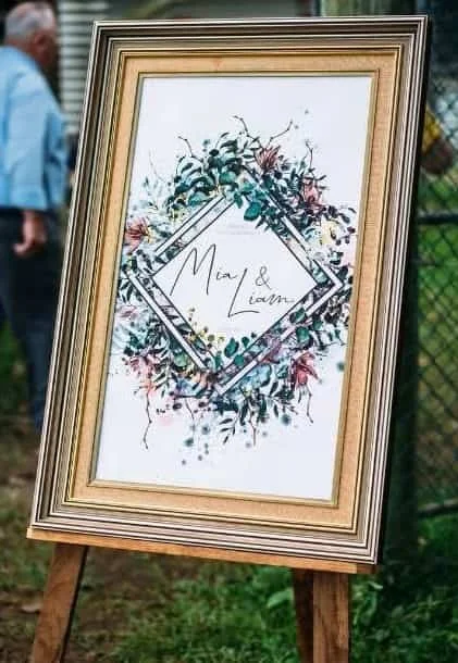 Decorative wedding sign with floral design and the names Mia and Liam in the center, displayed outdoors on an easel.
