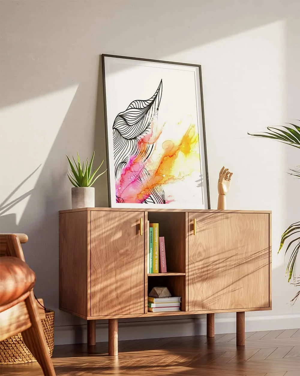 A wooden sideboard with yellow handles, decorated with a potted plant, a wooden mannequin hand, and a colorful abstract artwork leaning against the wall. Sunlight creates shadows on the wall and floor.