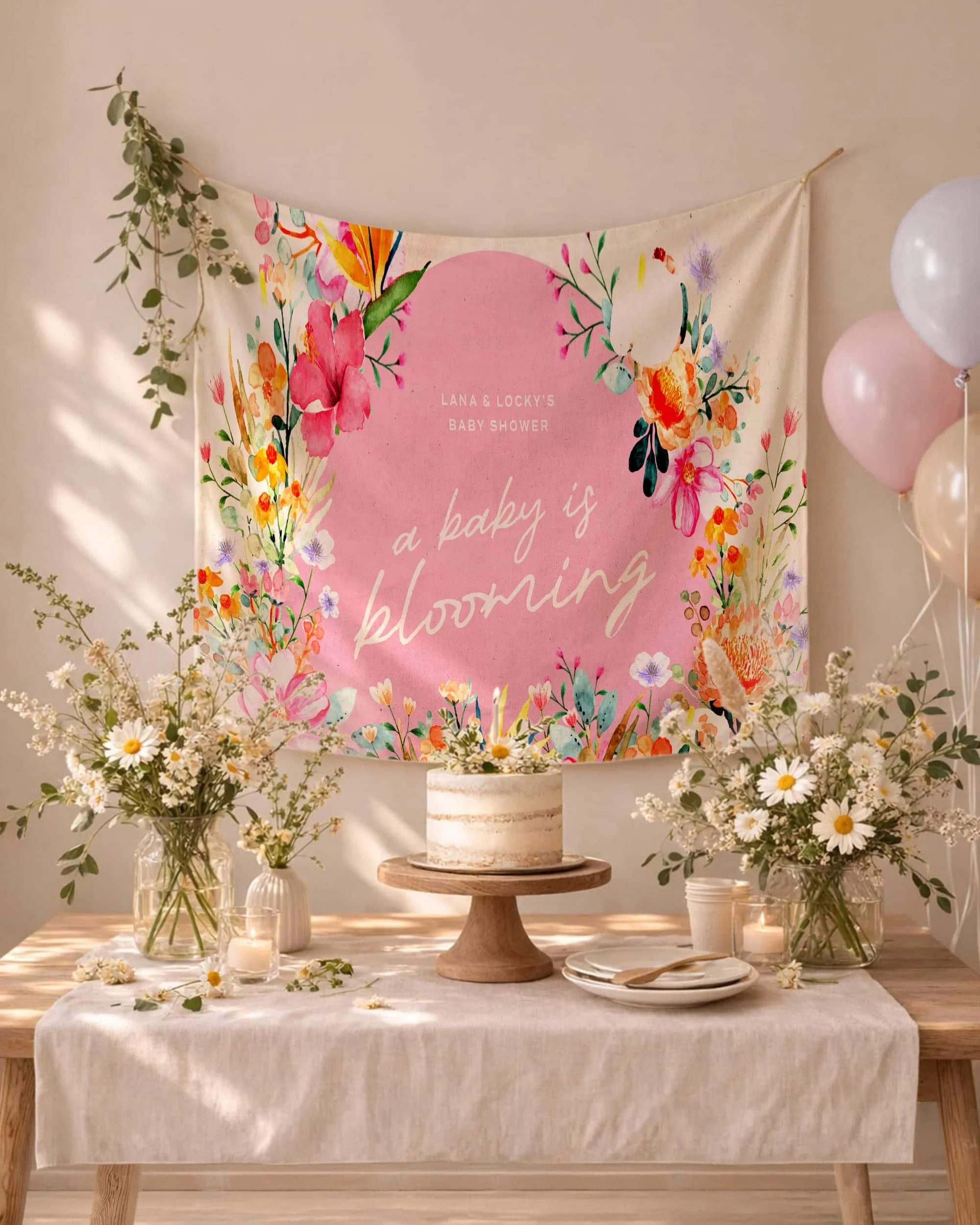 Decorative baby shower display with a pink and floral backdrop reading 'a baby is blooming,' flanked by white daisies in vases, candles, and a cake on a wooden table.