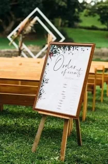 A framed wedding or event seating chart on an easel outdoors with wooden tables and chairs, and decorative geometric wall art in the background.
