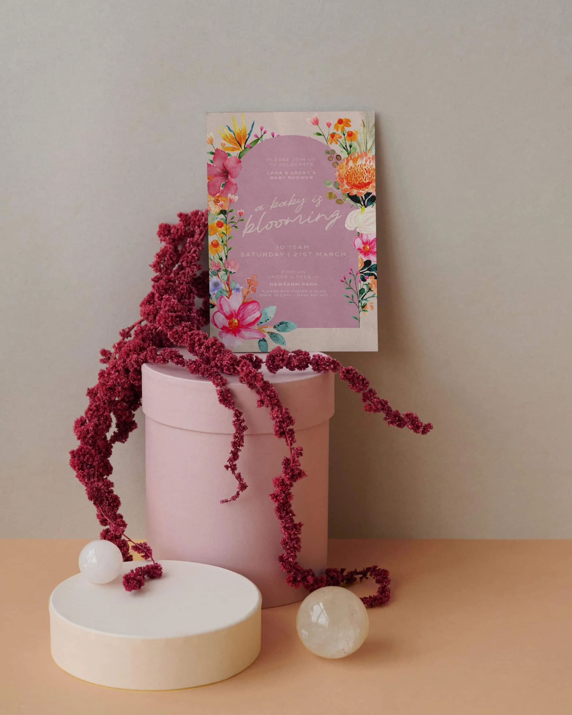 Pink flower pot with a pink floral baby shower invite, pink and white floral decorations, and a few decorative spheres on a wooden surface.
