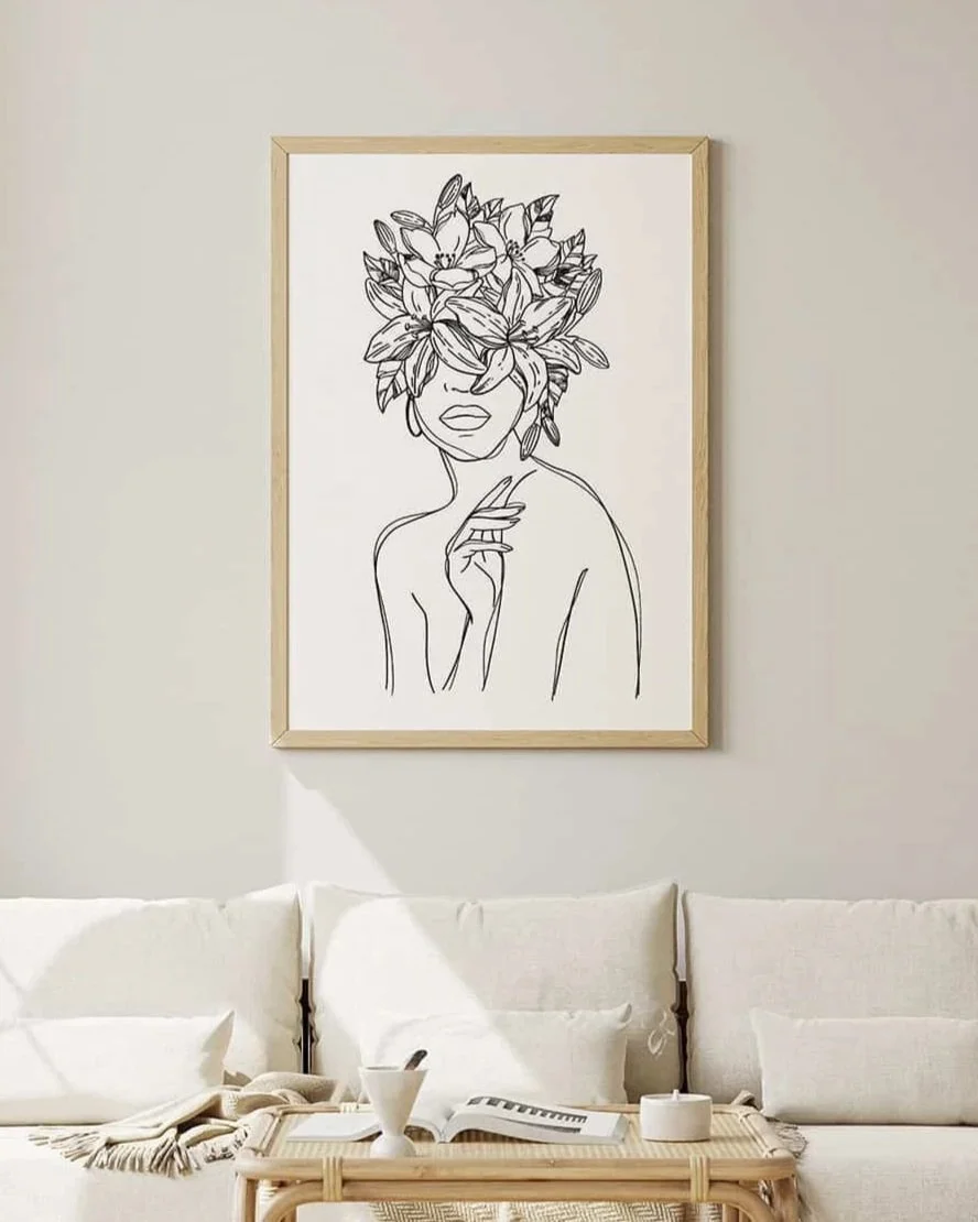 A modern minimalistic living room with a white sofa, pillows, a coffee table with a cup, an open book, and a notebook. Above the sofa, there is a framed line art drawing of a woman with large blooming flowers as her hair.