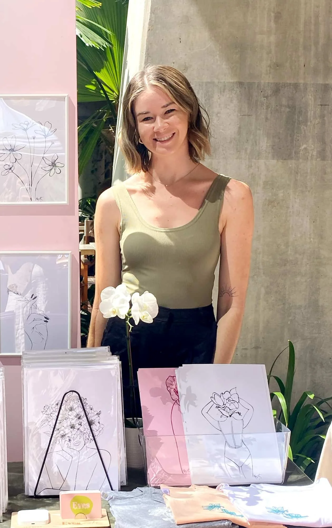 A smiling woman with shoulder-length hair standing behind a display table with art prints, a white orchid, and clothing on a table. The background includes a gray wall, a pink panel with line art drawings, and tropical plants.