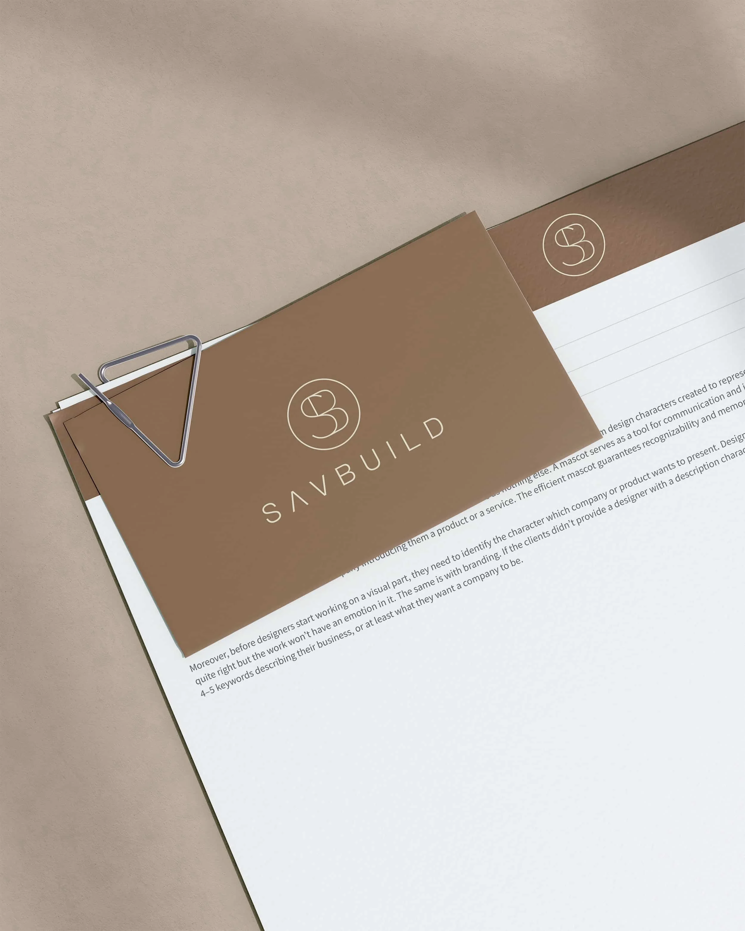 Brown business card with white logo and text that says 'SAVBUILD', clipped to a white sheet of paper with printed text, held by a silver paperclip on beige background.