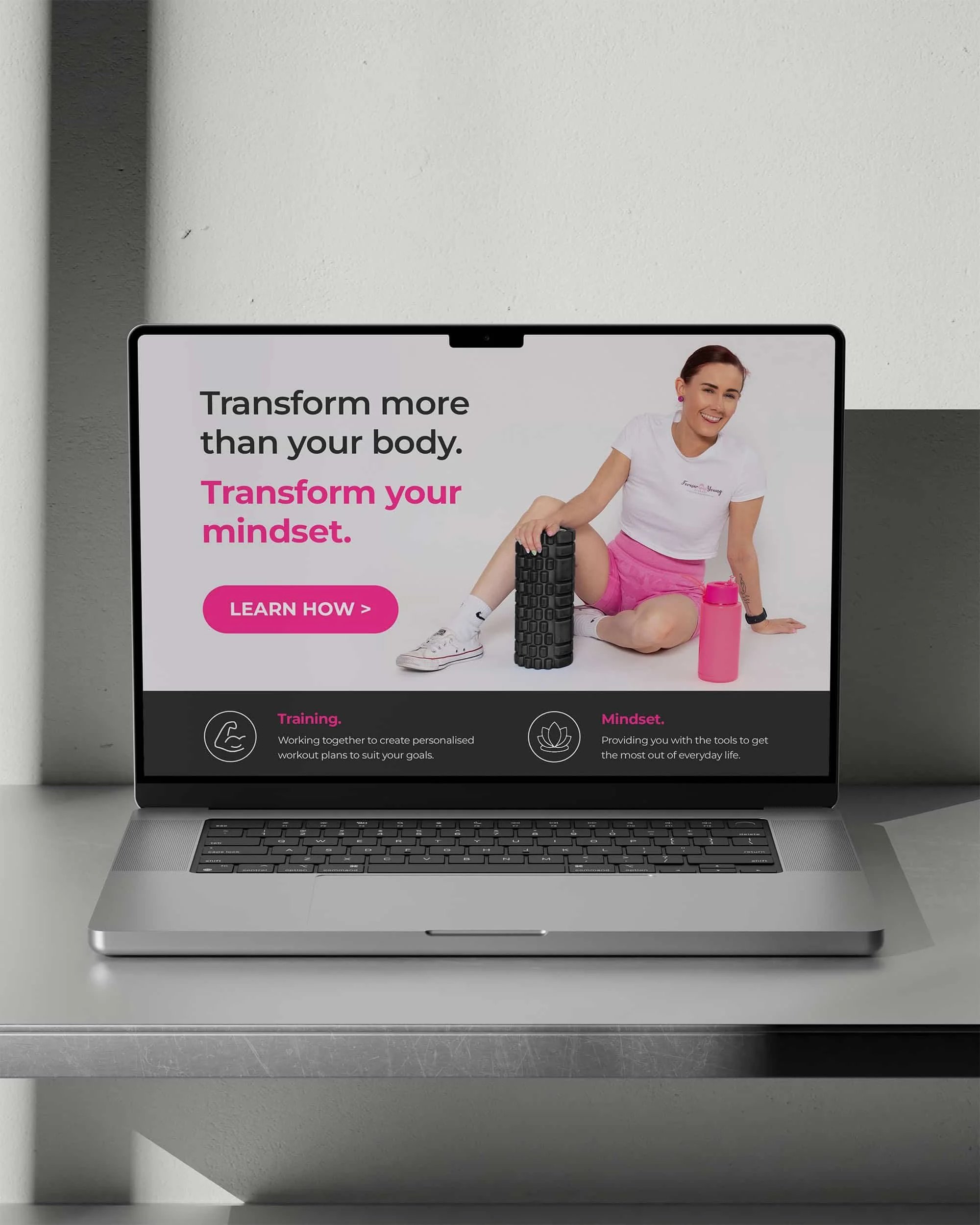 Laptop screen displaying an advertisement for fitness and mindset coaching, with a smiling woman in athletic clothing sitting next to a foam roller and water bottle.