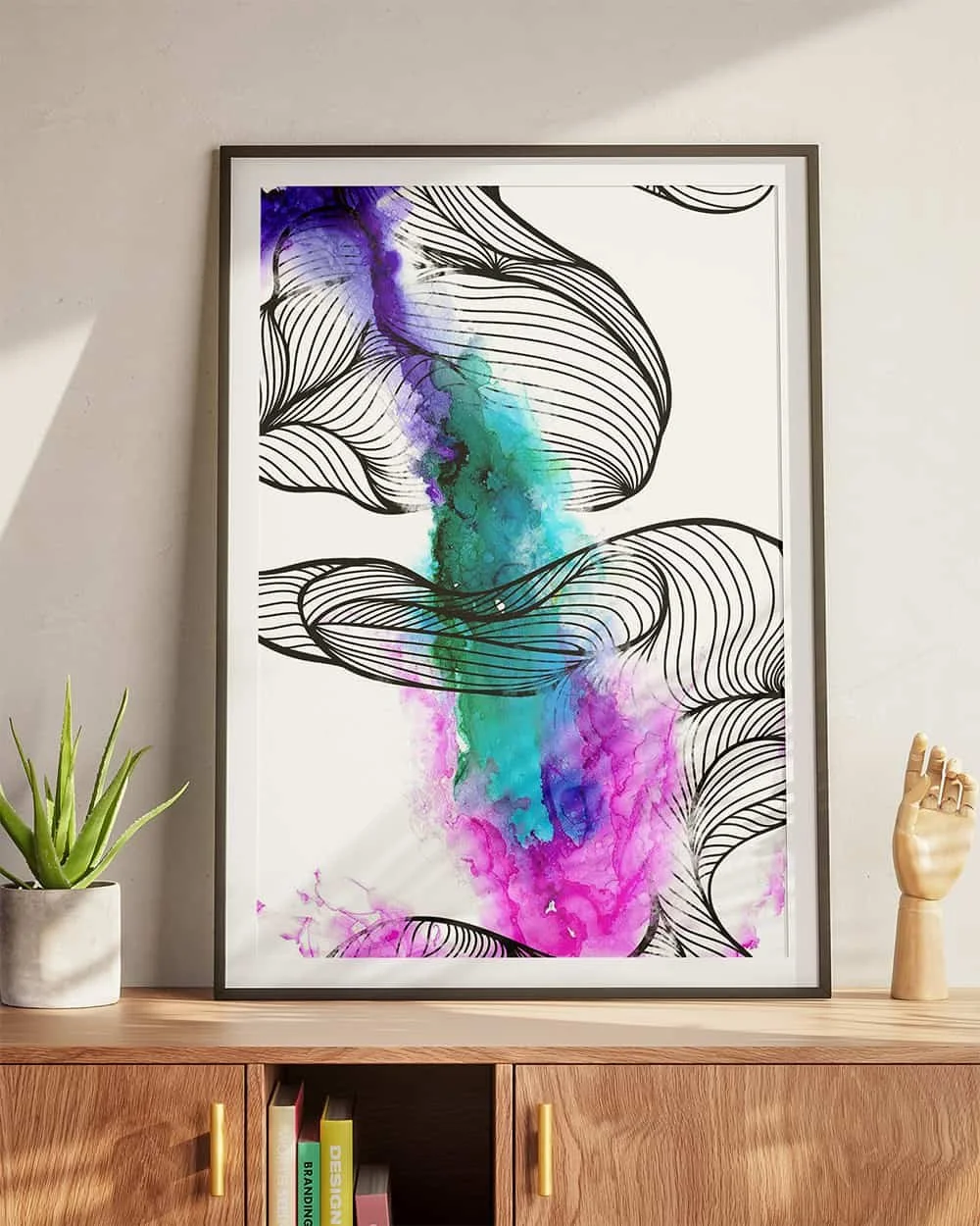 Framed abstract art print with black line art and colorful watercolor splashes, on a wooden sideboard next to potted plant and mannequin hand.