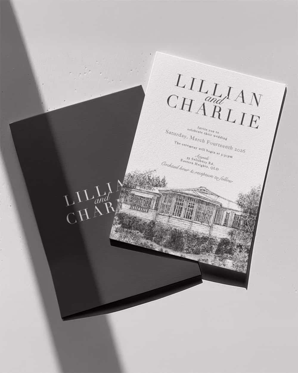 Wedding invitation card with house illustration and text for Lillian and Charlie's wedding, next to a matching black envelope, on a white surface.