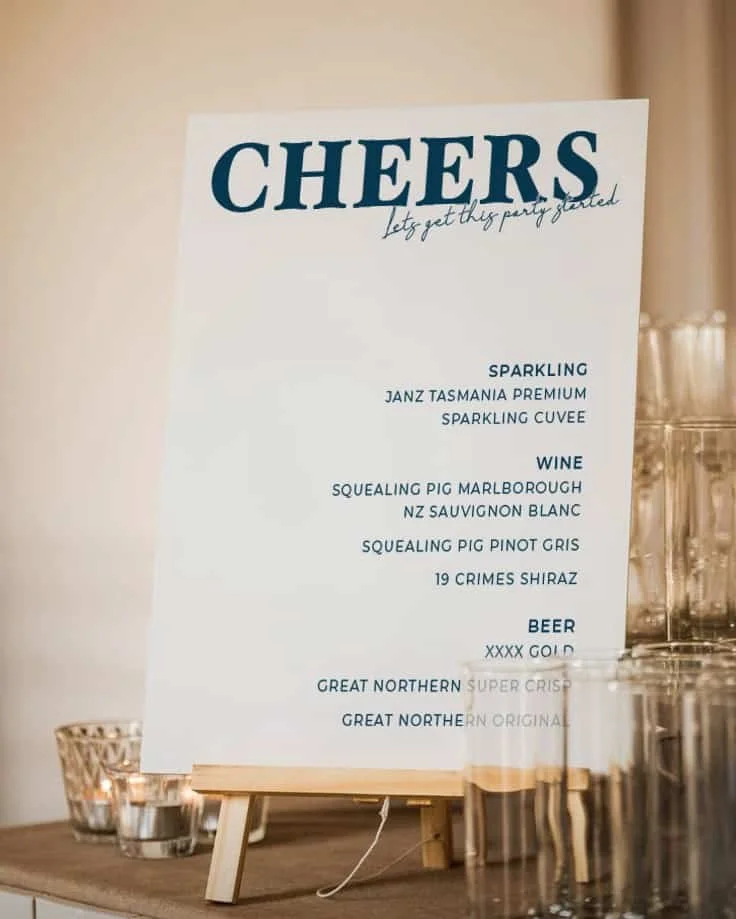 Party beverage menu on a white easel listing options such as sparkling wine, NZ Sauvignon Blanc, Pinot Gris, Shiraz, and various beers, with glasses and candles on a wooden table nearby.